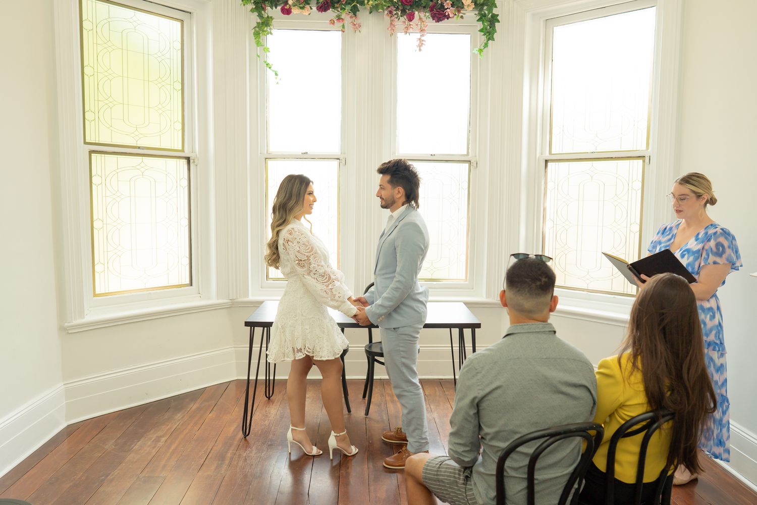 Small intimate wedding ceremony takes place in bright corner room with bay windows and floral decorations.