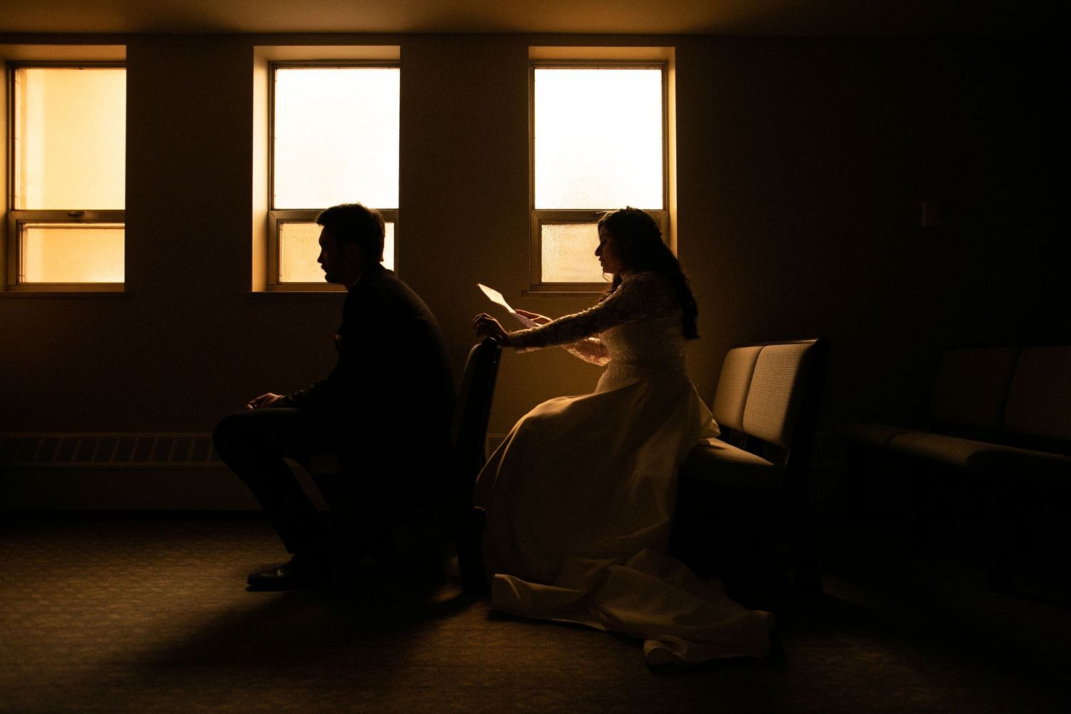 Two silhouettes seated by bright windows in dark moody room.