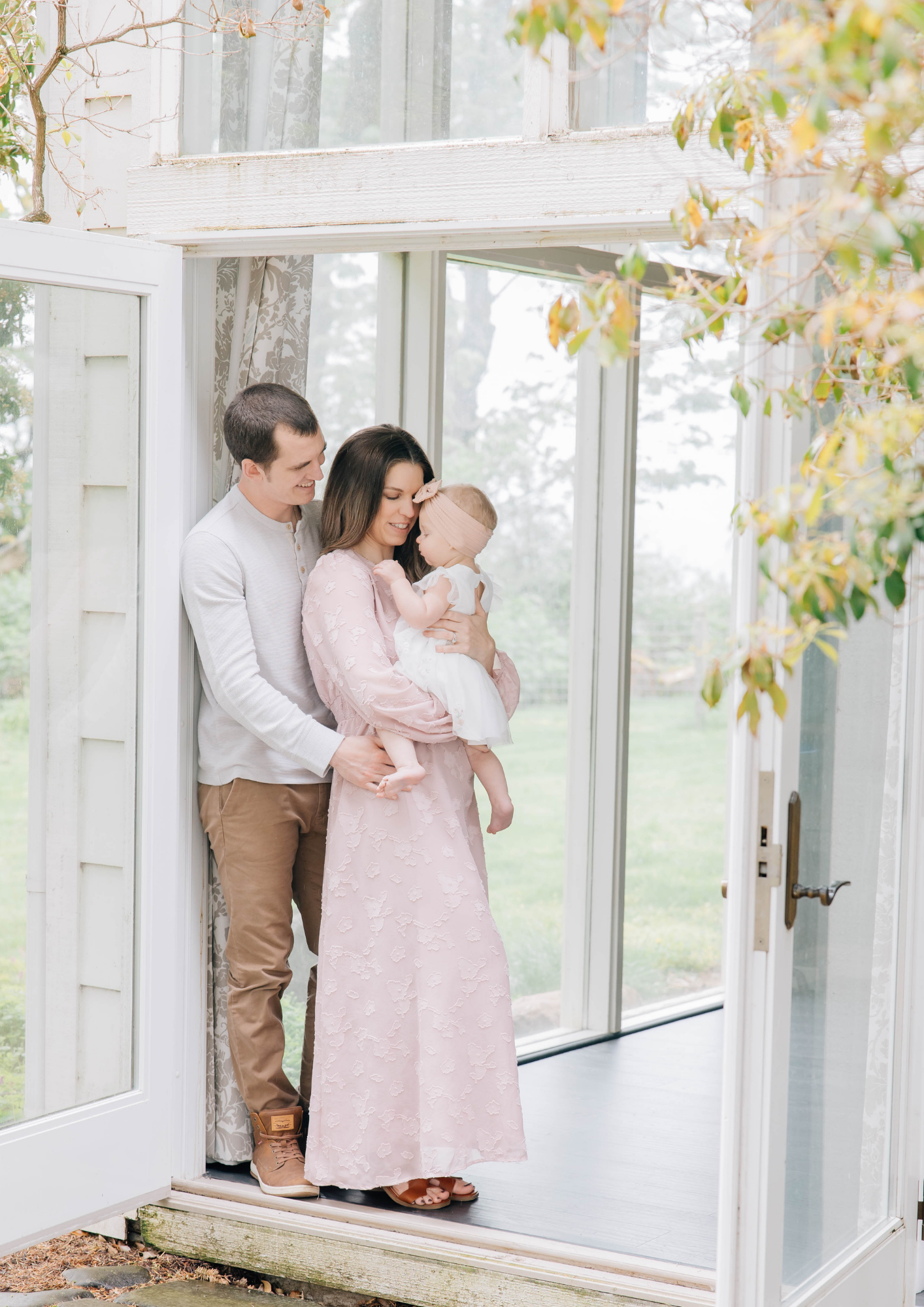 A couple tenderly holds their baby while standing in an elegant white doorway framed by autumn leaves.