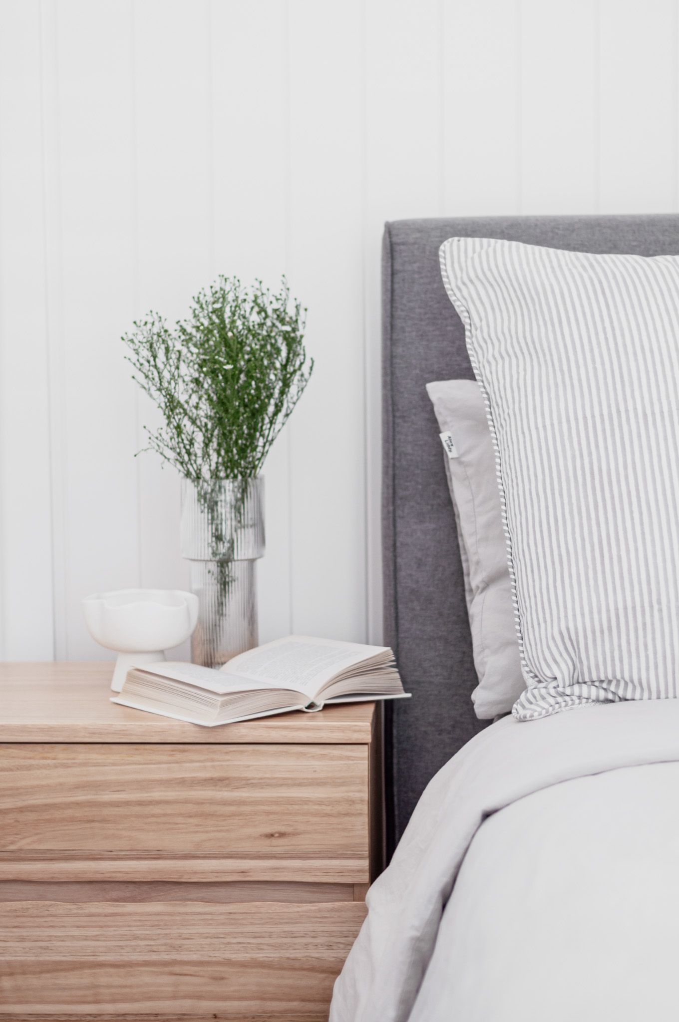 Serene bedside scene with fresh greenery, open book, and striped pillows on gray headboard.