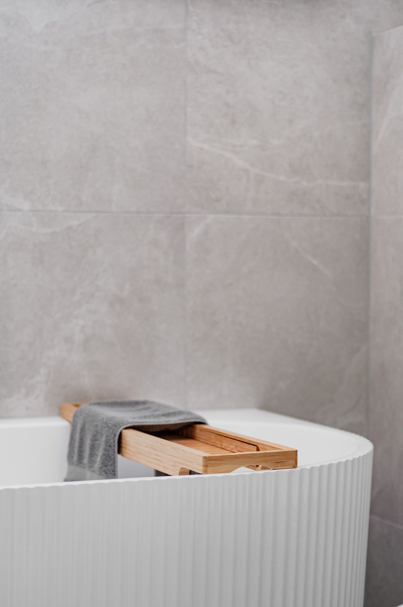 Luxurious bathtub with wooden bath caddy and gray towel against marble-look tile walls.