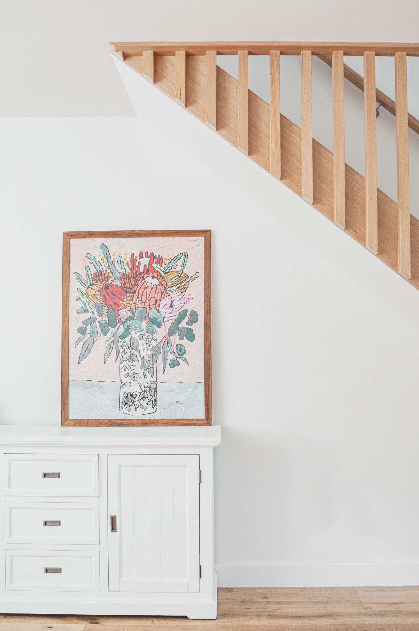 Wooden staircase with white walls and floral artwork displayed on white cabinet below.