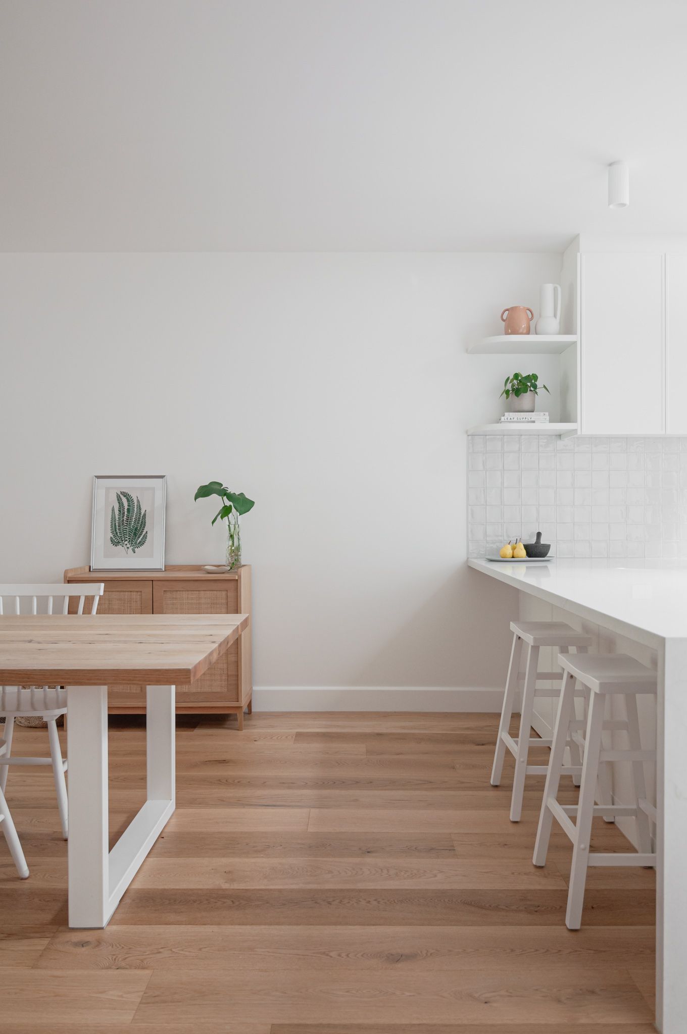 Minimalist dining area with wooden table, white chairs, and botanical artwork against white walls.