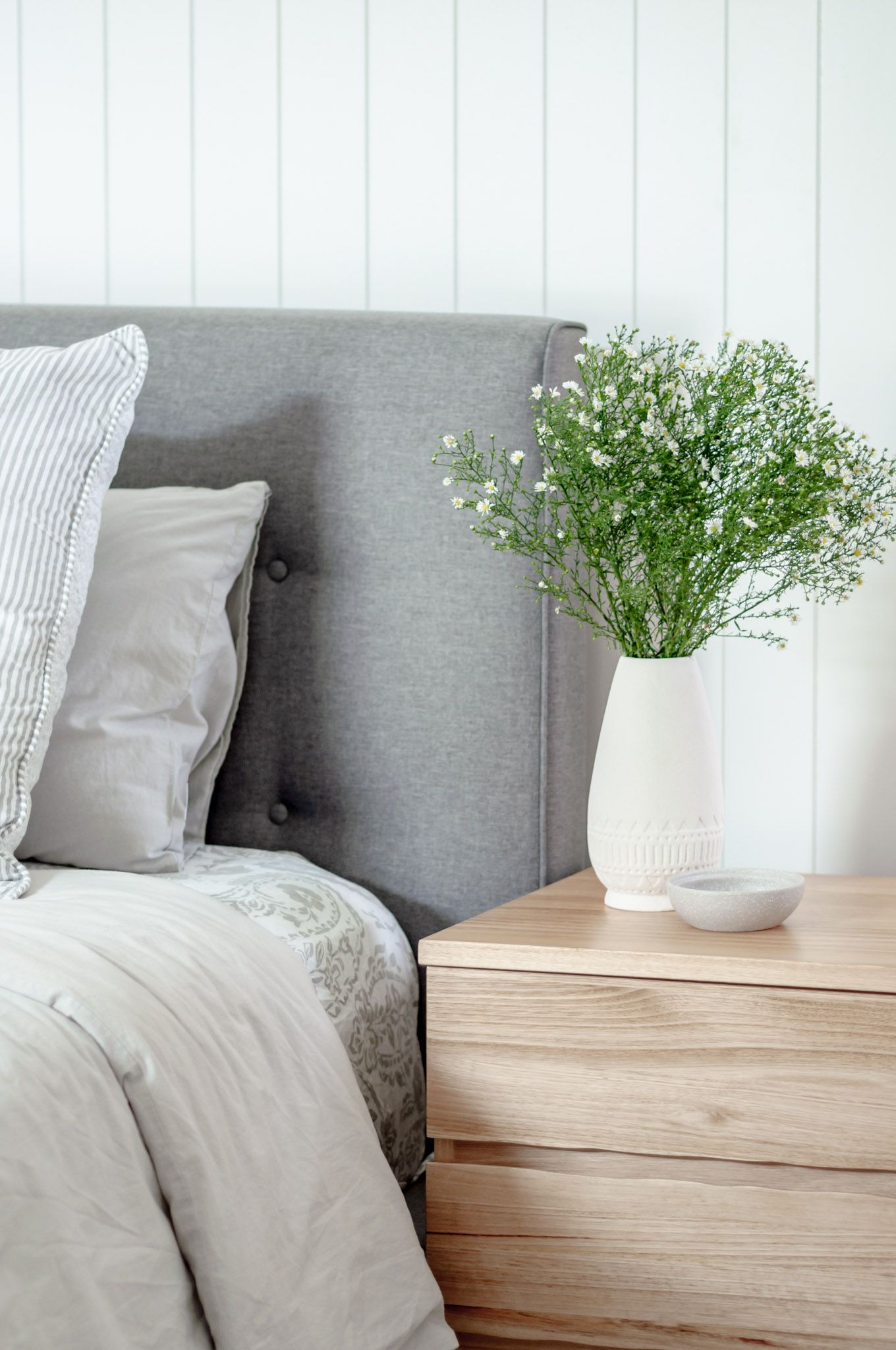 Modern bedside table with white vase of fresh flowers next to gray upholstered headboard.