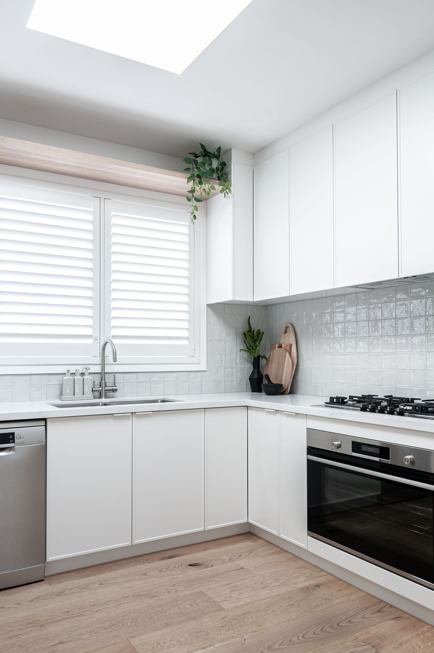 White modern kitchen with wood flooring, plantation shutters, and hanging plants near window.