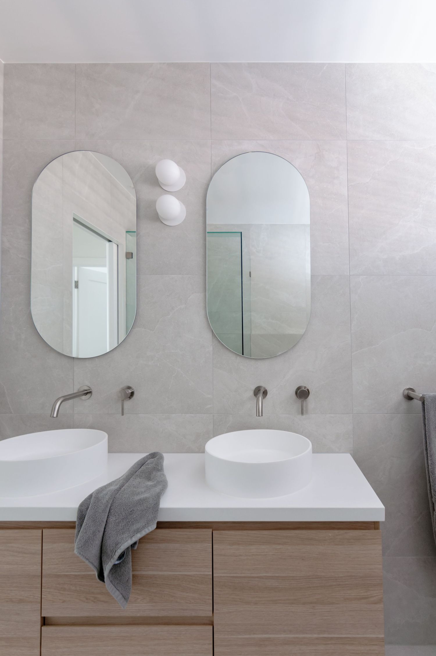 Modern bathroom interior with two oval mirrors, white vessel sinks, and wooden vanity against light gray tile walls.
