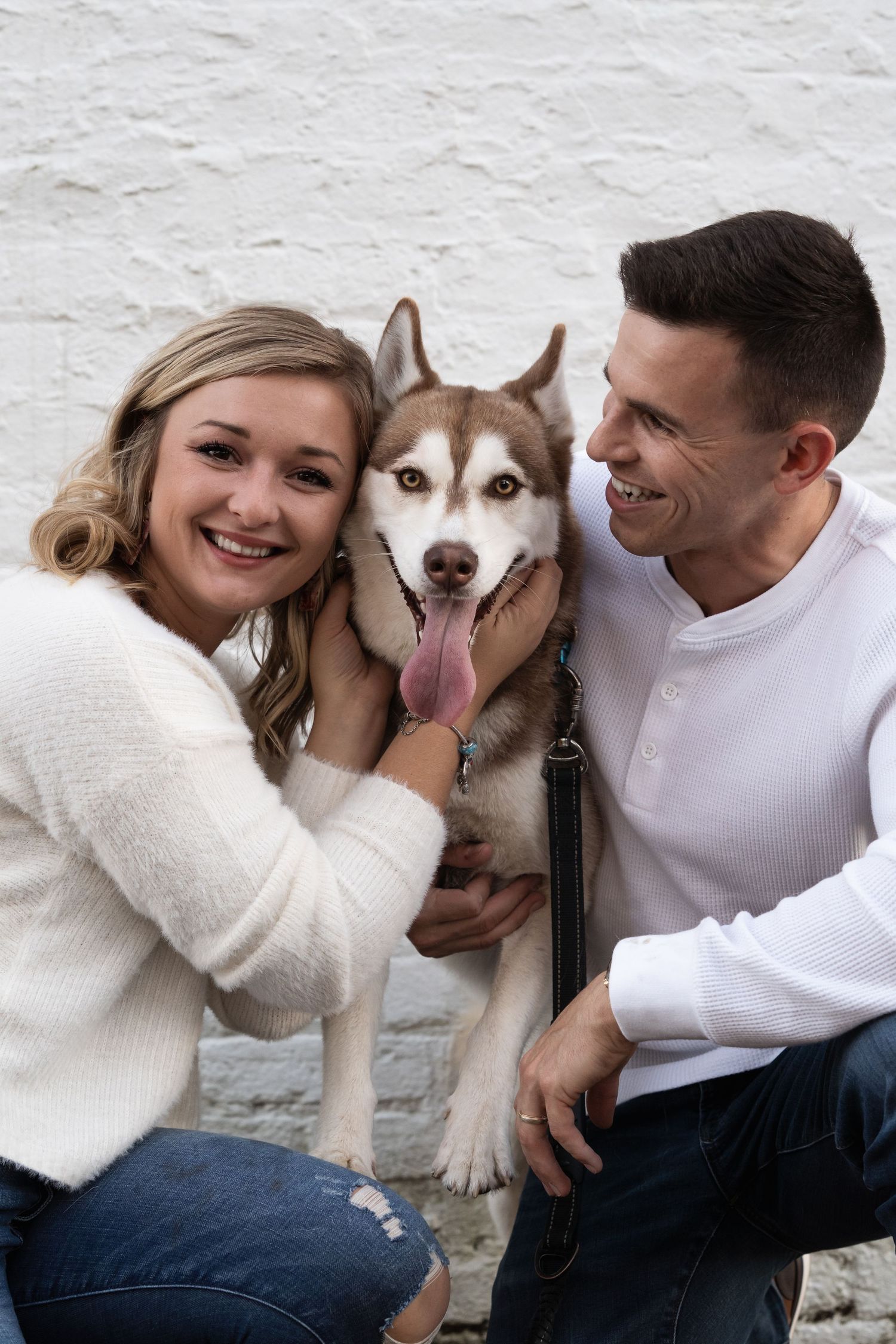 A happy couple poses with their smiling Husky dog against a white wall.