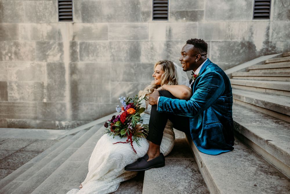 Newly married couple sits on stone steps with vibrant bouquet in urban setting.