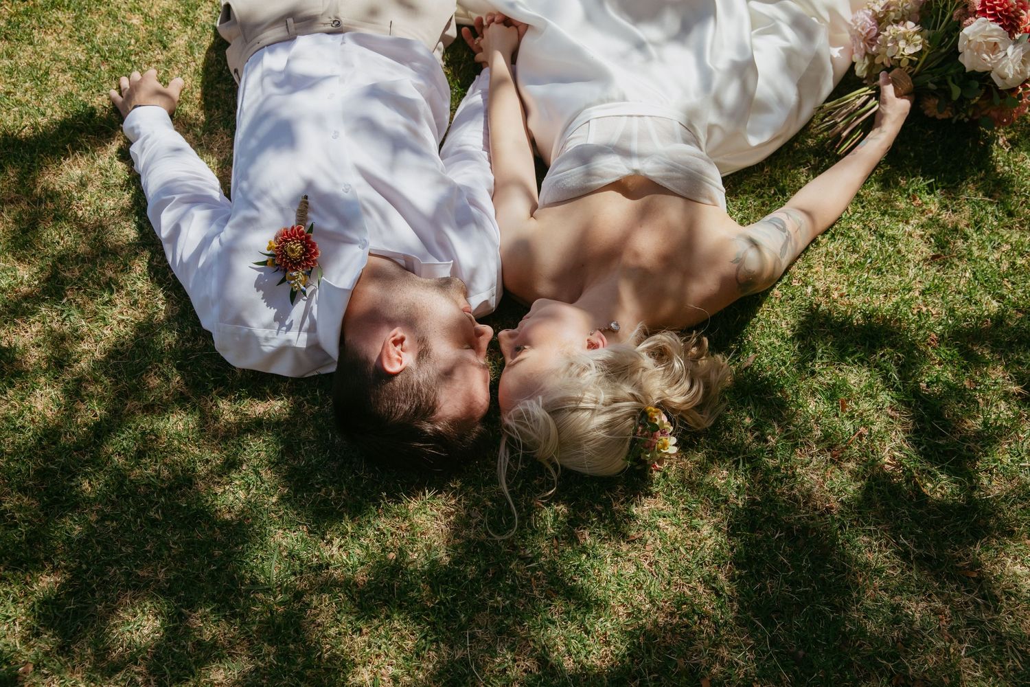 Couple lying on grass in wedding attire with dappled sunlight and flowers around them.