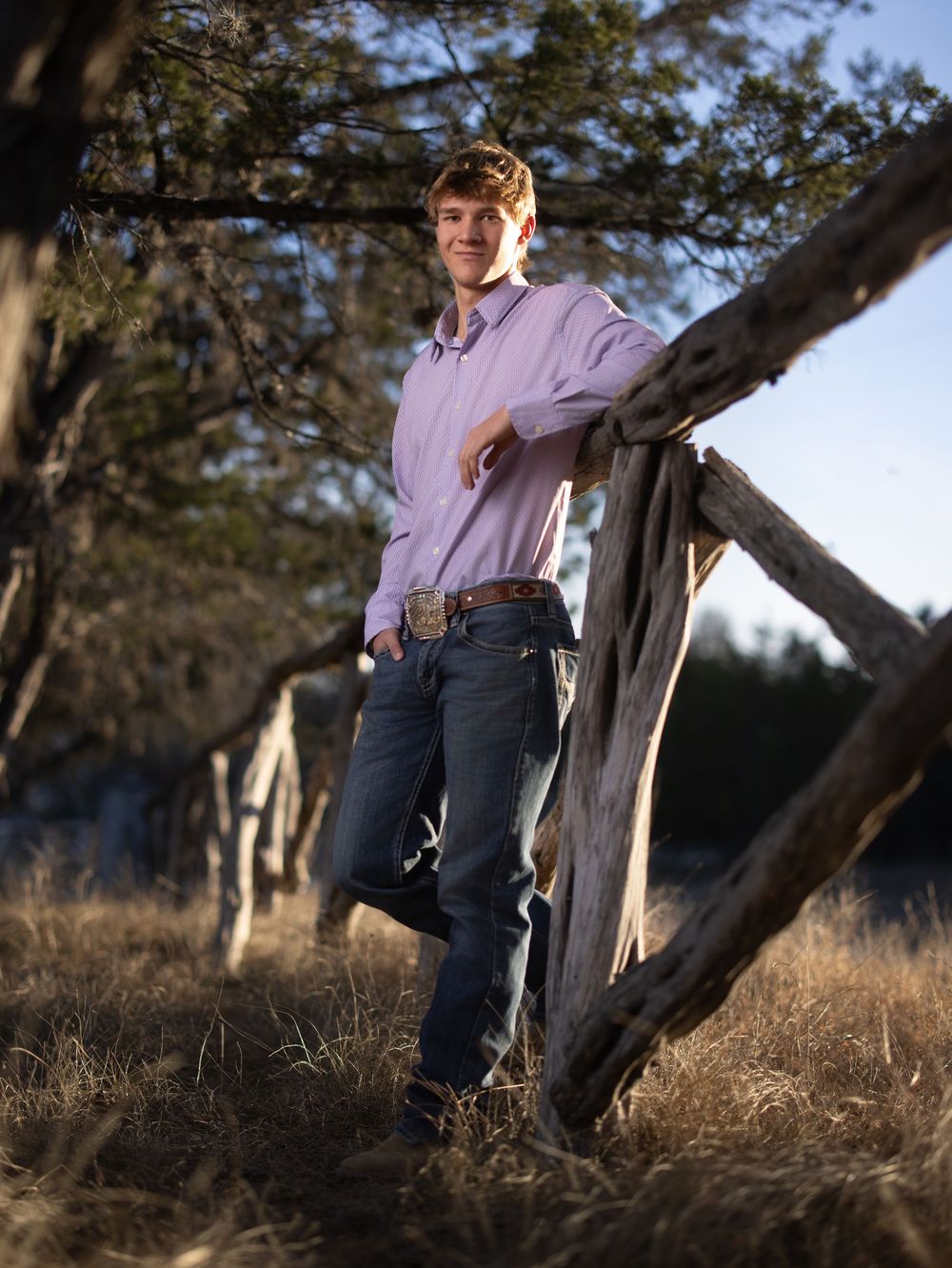 Young man in a purple button-down shirt and jeans poses casually against a rustic tree trunk in natural outdoor lighting.
