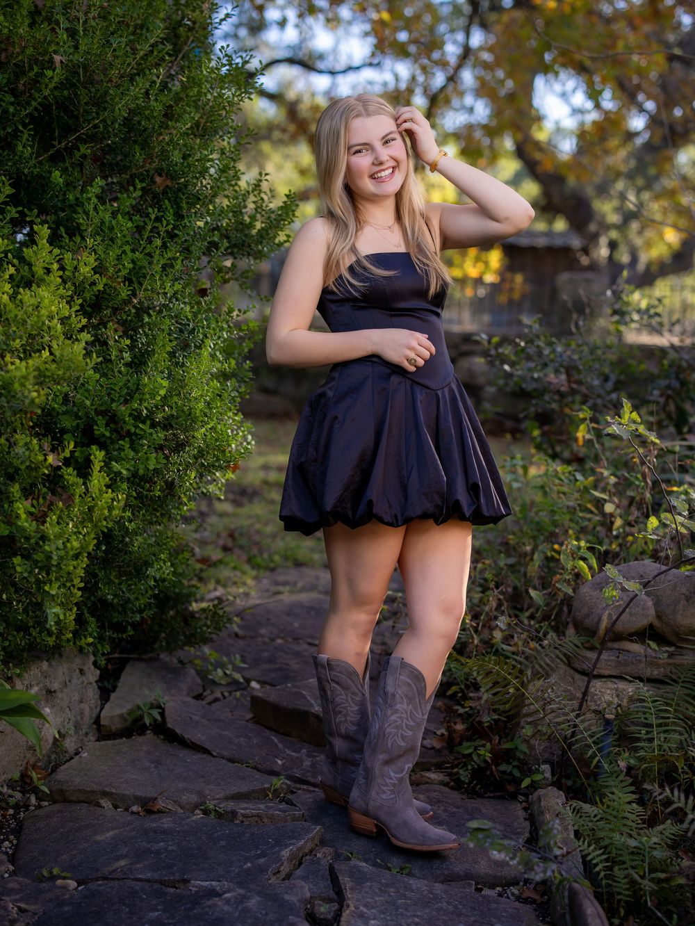 A teenager in a black dress and brown cowboy boots poses outdoors on rocky terrain surrounded by autumn foliage.