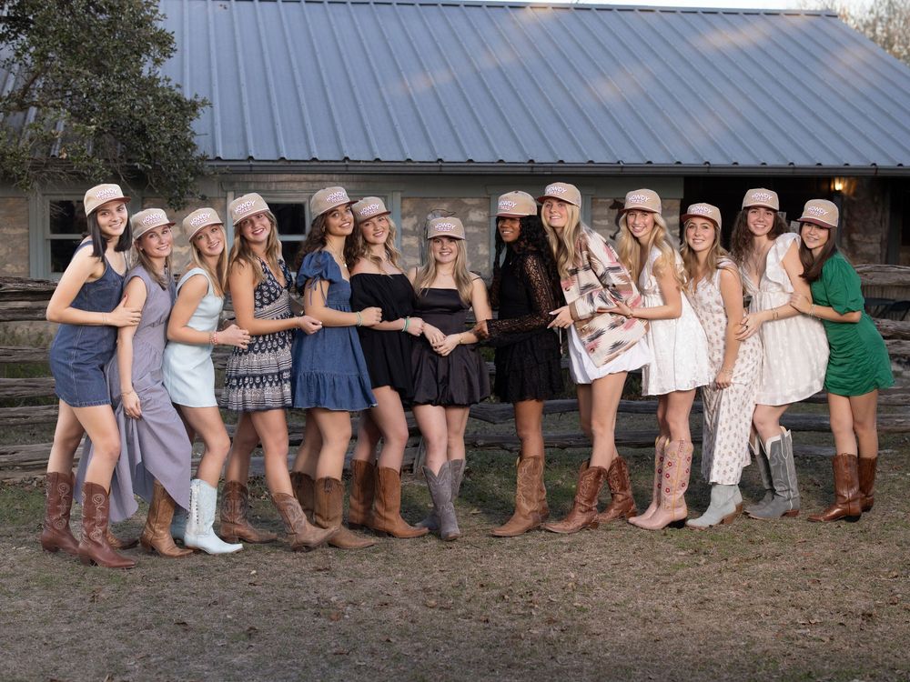 A row of people wearing boots and cowboy boots stand together showing off their footwear against a rustic backdrop.
