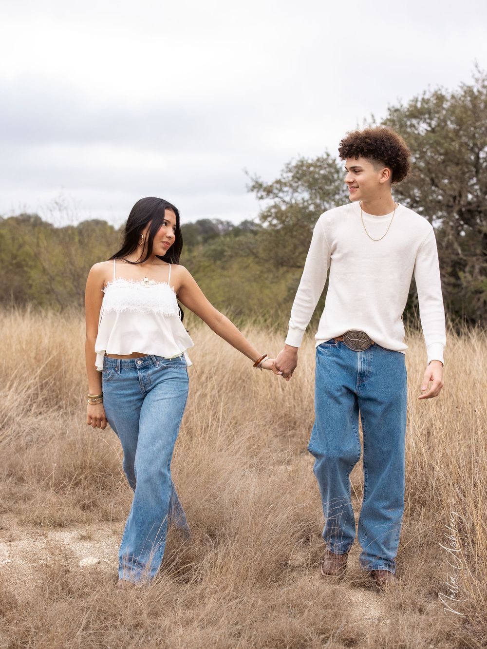 Young couple holding hands while standing in a golden grass field on an overcast day wearing jeans and casual attire.