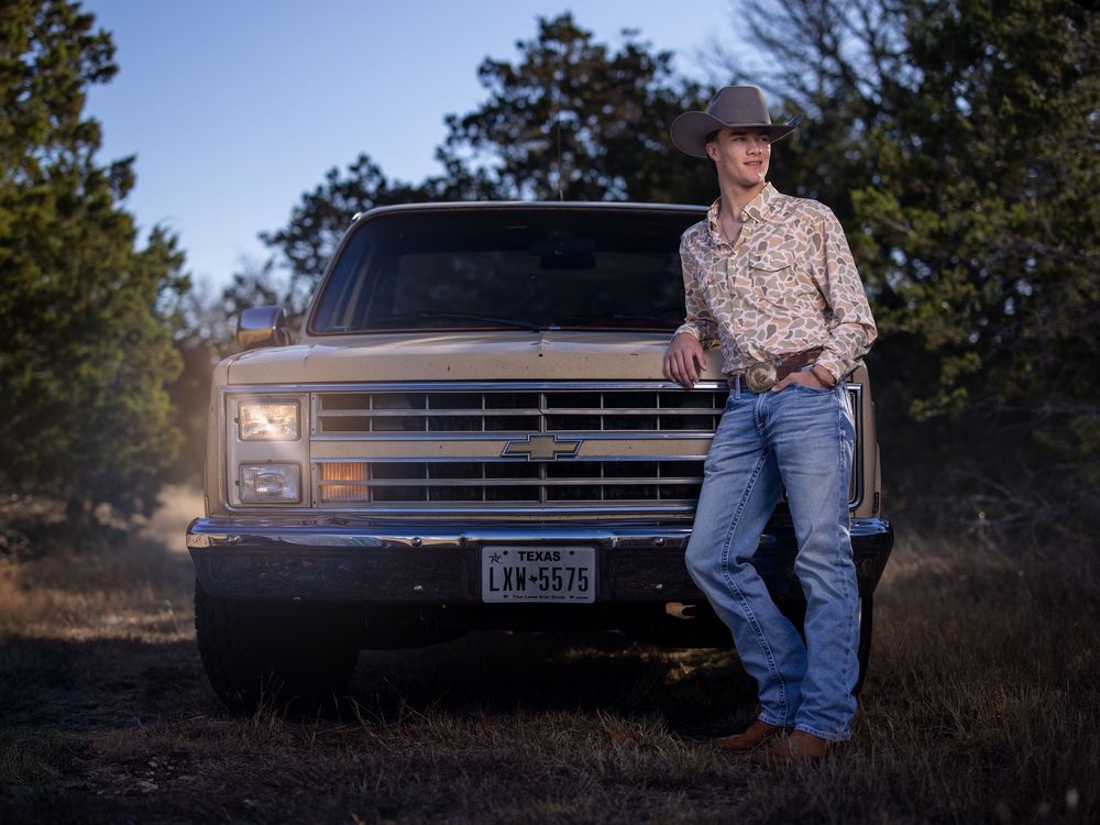 A classic Chevrolet pickup truck parked in a wooded area at dusk with a western-styled young manstanding beside it.