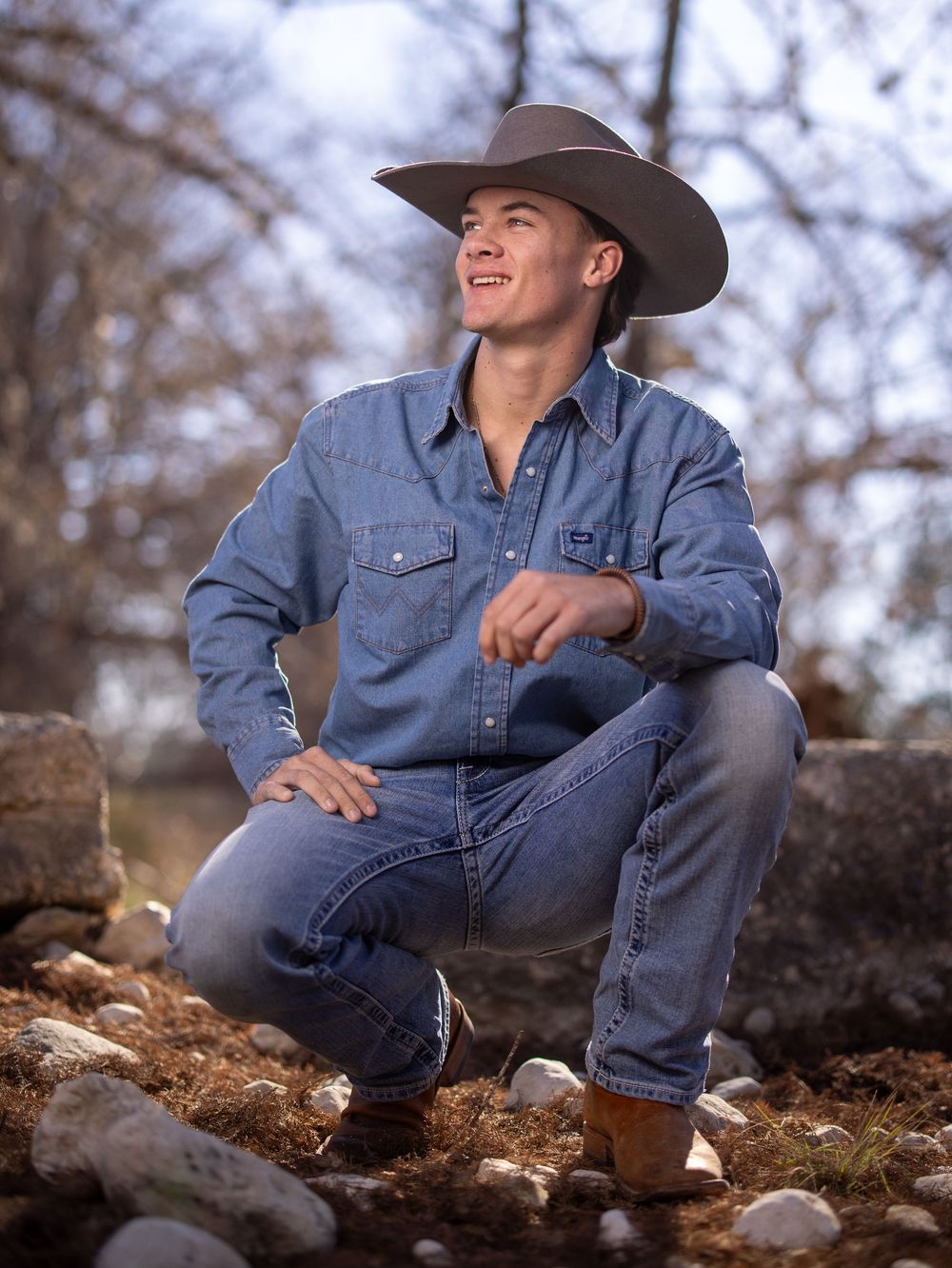 A young man in denim and cowboy hat sitting casually on rocky terrain against a blurred natural backdrop.