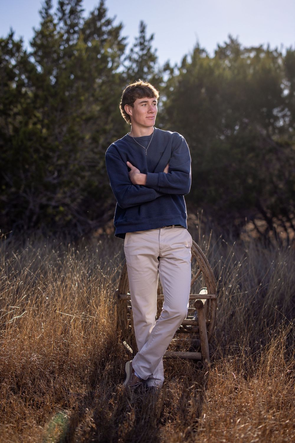 A young man in navy sweater and khaki pants poses thoughtfully in a field of tall dried grass at golden hour.
