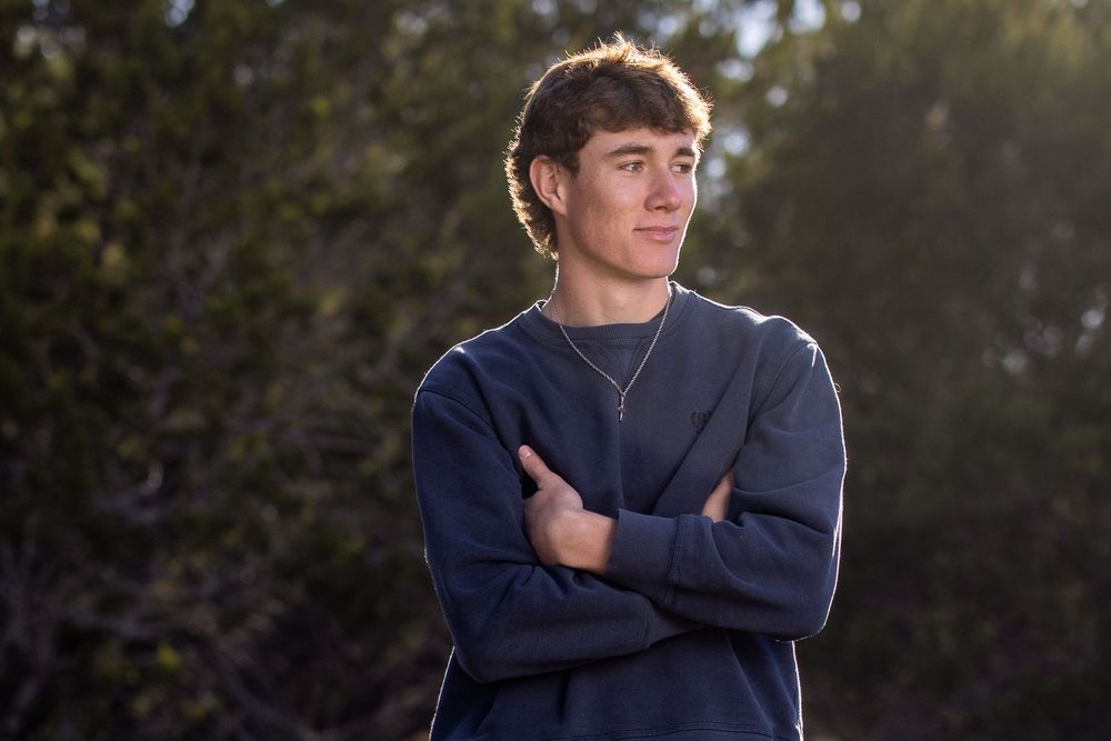 A young man in a navy blue sweater stands with arms crossed outdoors during golden hour lighting.