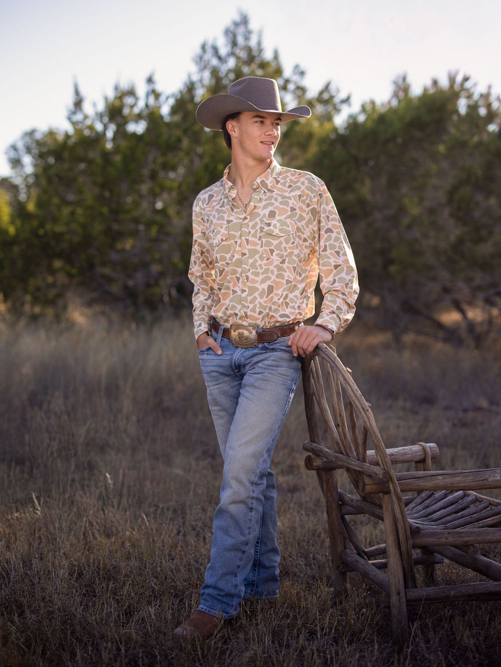 A young man in jeans and Burlebo top leans casually against a rustic wooden fence in a rural field setting at dusk.
