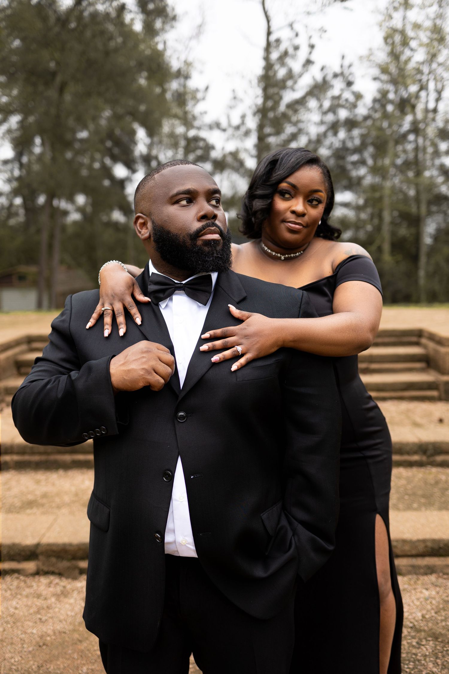 Elegant couple in formal black attire pose together during an outdoor photoshoot.