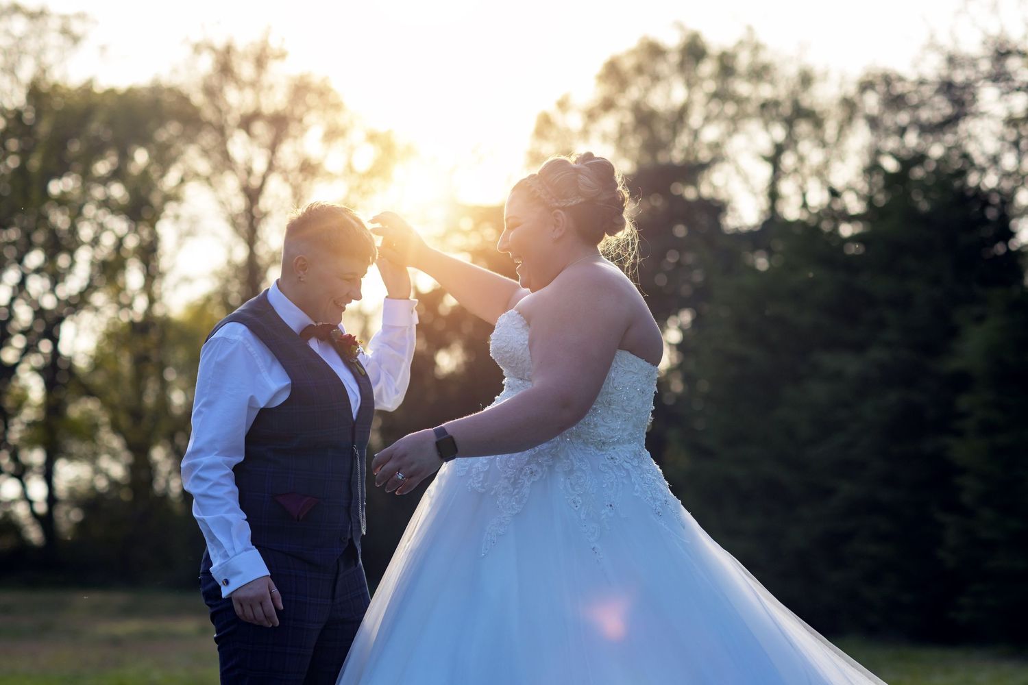 Same-sex couple dancing outdoors at sunset, with one partner in a white wedding gown and the other in a vest and shirt