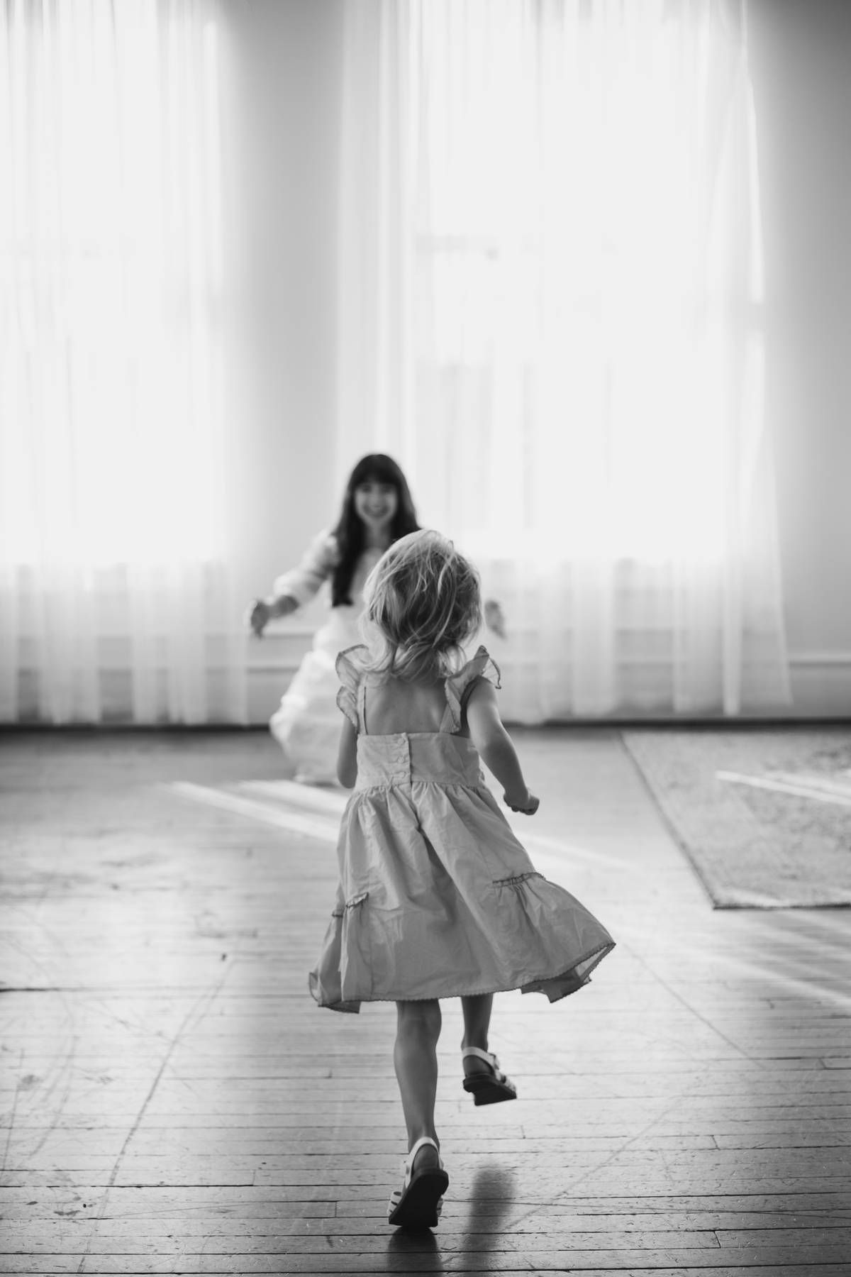 Black and white photo of a little girl in a dress running across a wood floor toward a smiling woman with open arms.