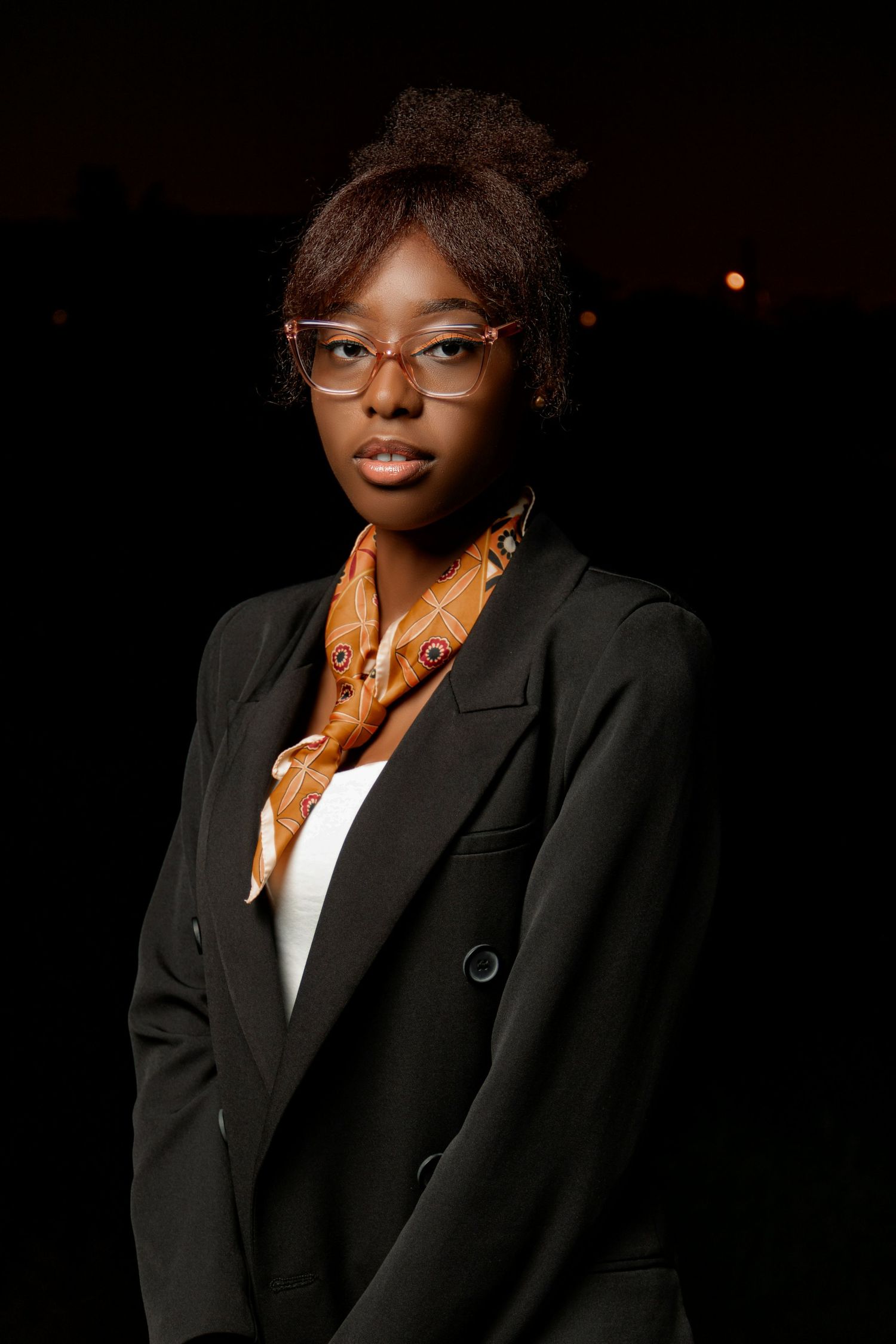 Professional portrait of someone in a black blazer and patterned scarf wearing glasses.