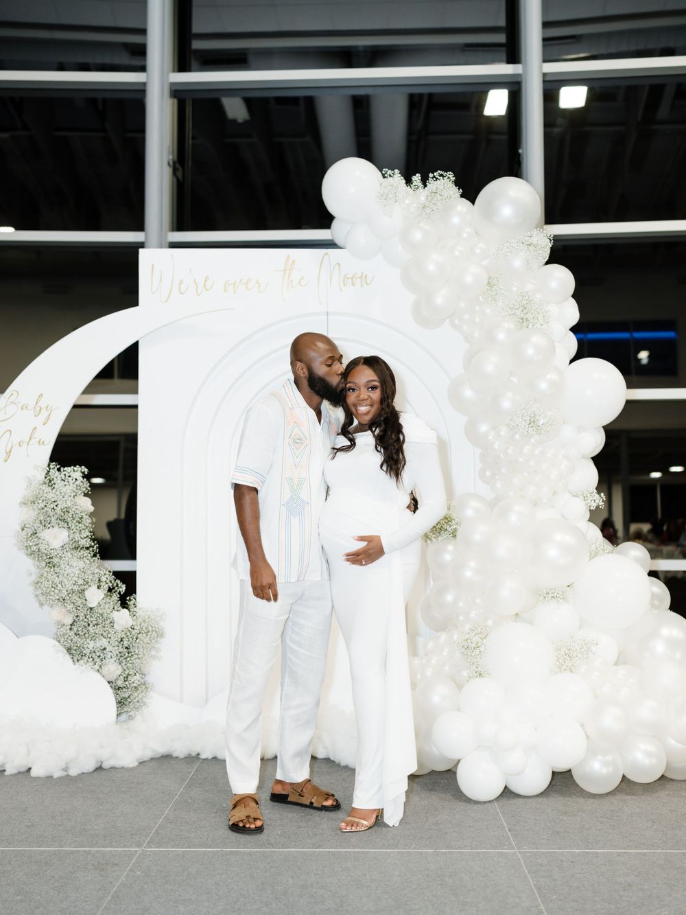 A couple in white outfits poses in front of an elegant white balloon arch and floral backdrop.