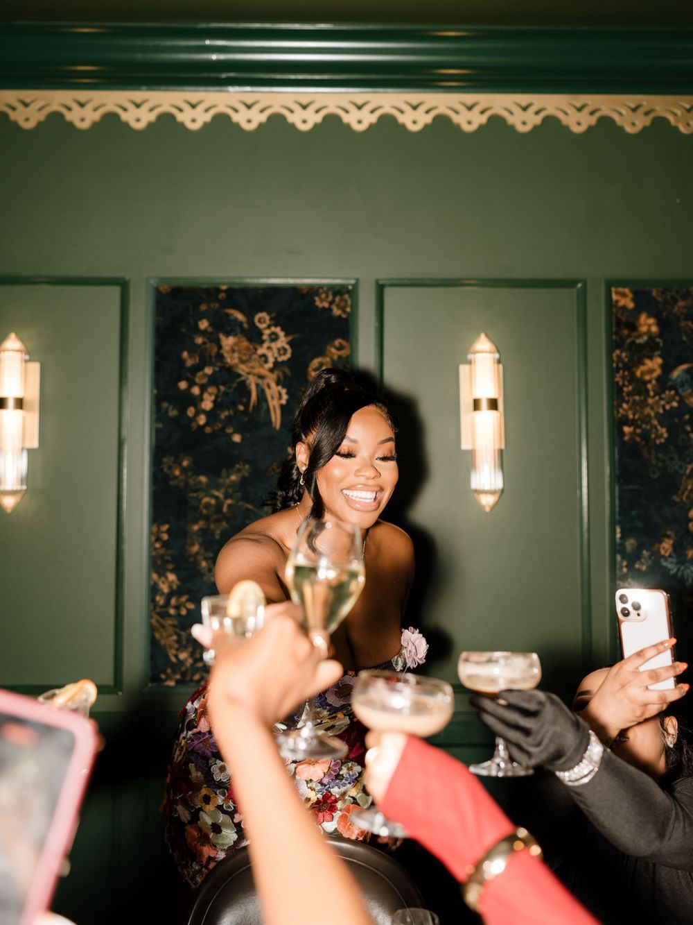 Friends raise champagne glasses in a toast at an elegant celebration with wall sconces and floral wallpaper.