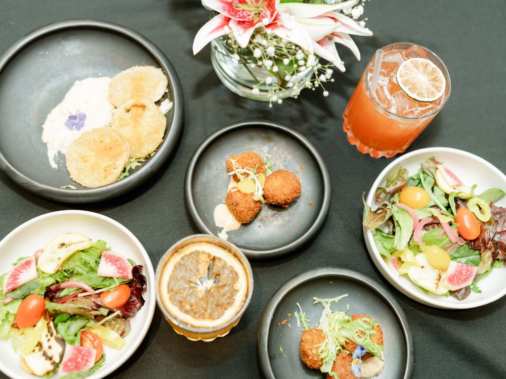 Overhead view of colorful appetizers and cocktails served on dark plates with a floral centerpiece.