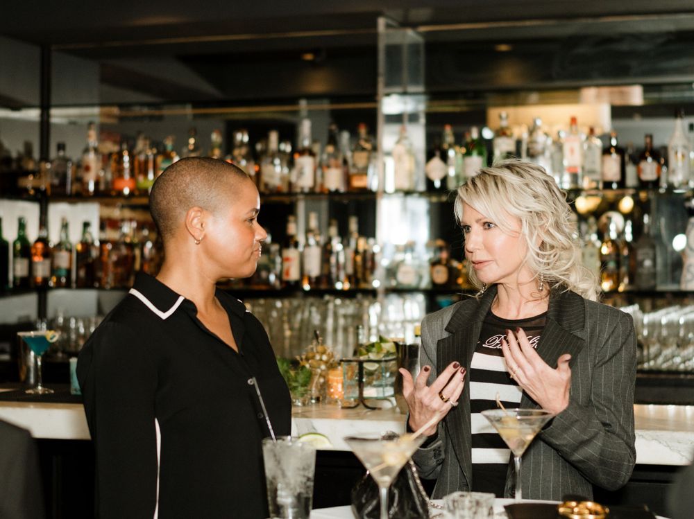 Two people having an animated conversation while standing at a bar with shelves of bottles in the background.