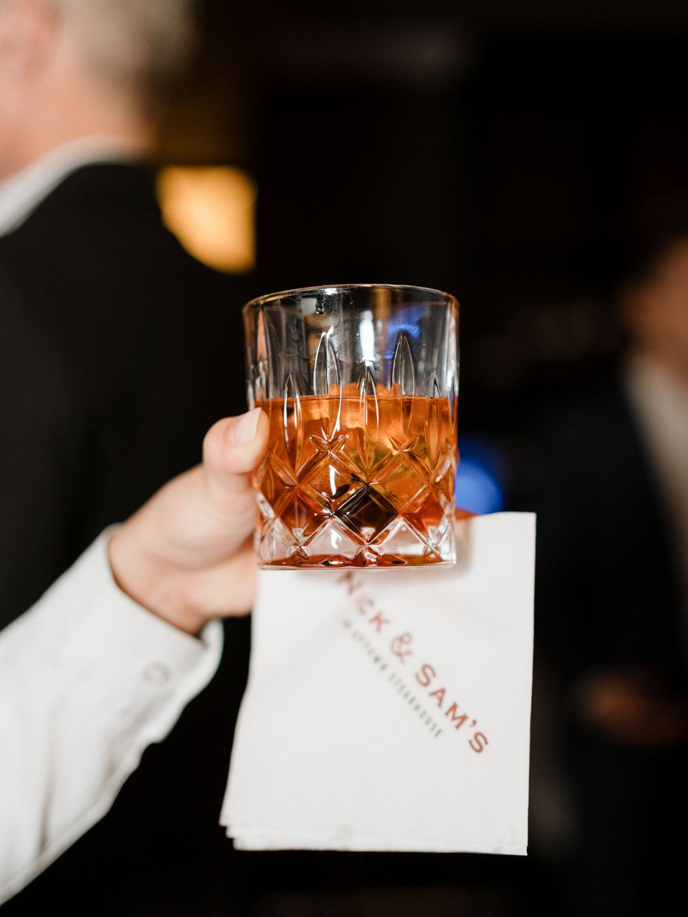 A crystal cocktail glass filled with amber-colored spirit is held against a white napkin in a dimly lit setting.