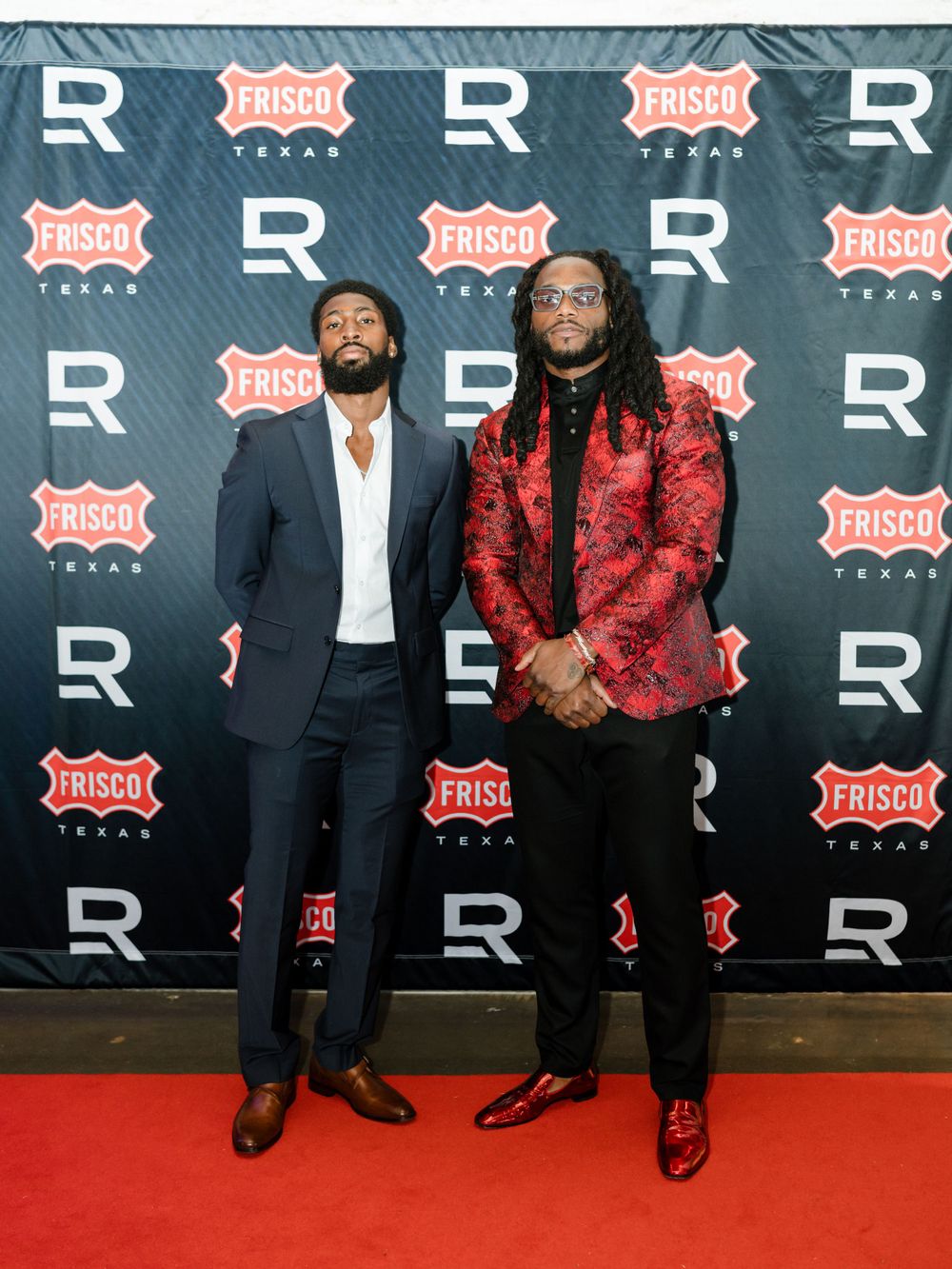 Two men in formal attire pose on red carpet against backdrop with R and Frisco branding.
