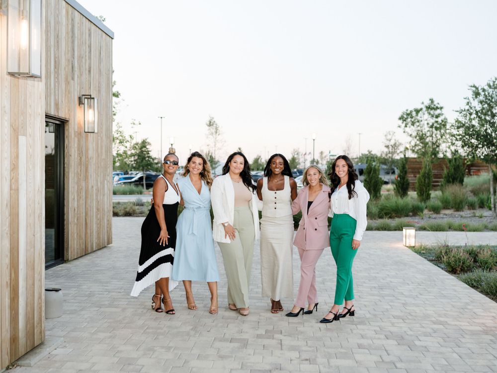 Group of women in coordinated pastel outfits pose together on stone pathway outside modern building.