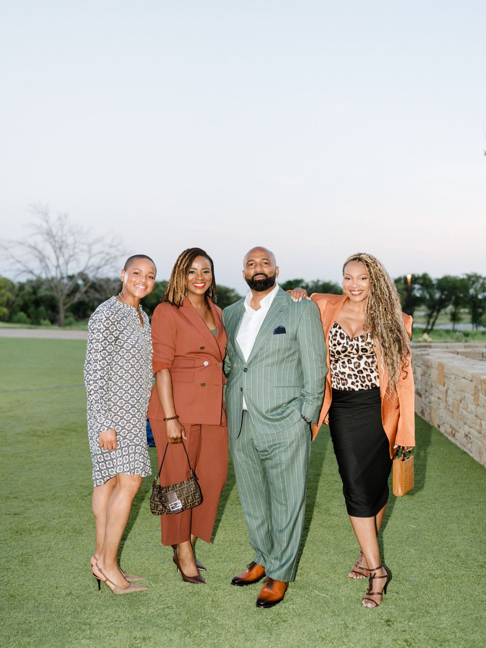 Group of four people in business attire pose together on grass with sunset backdrop.