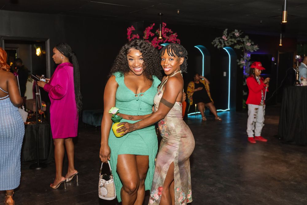 Two partygoers in festive dresses pose together at a dimly lit nightclub event.