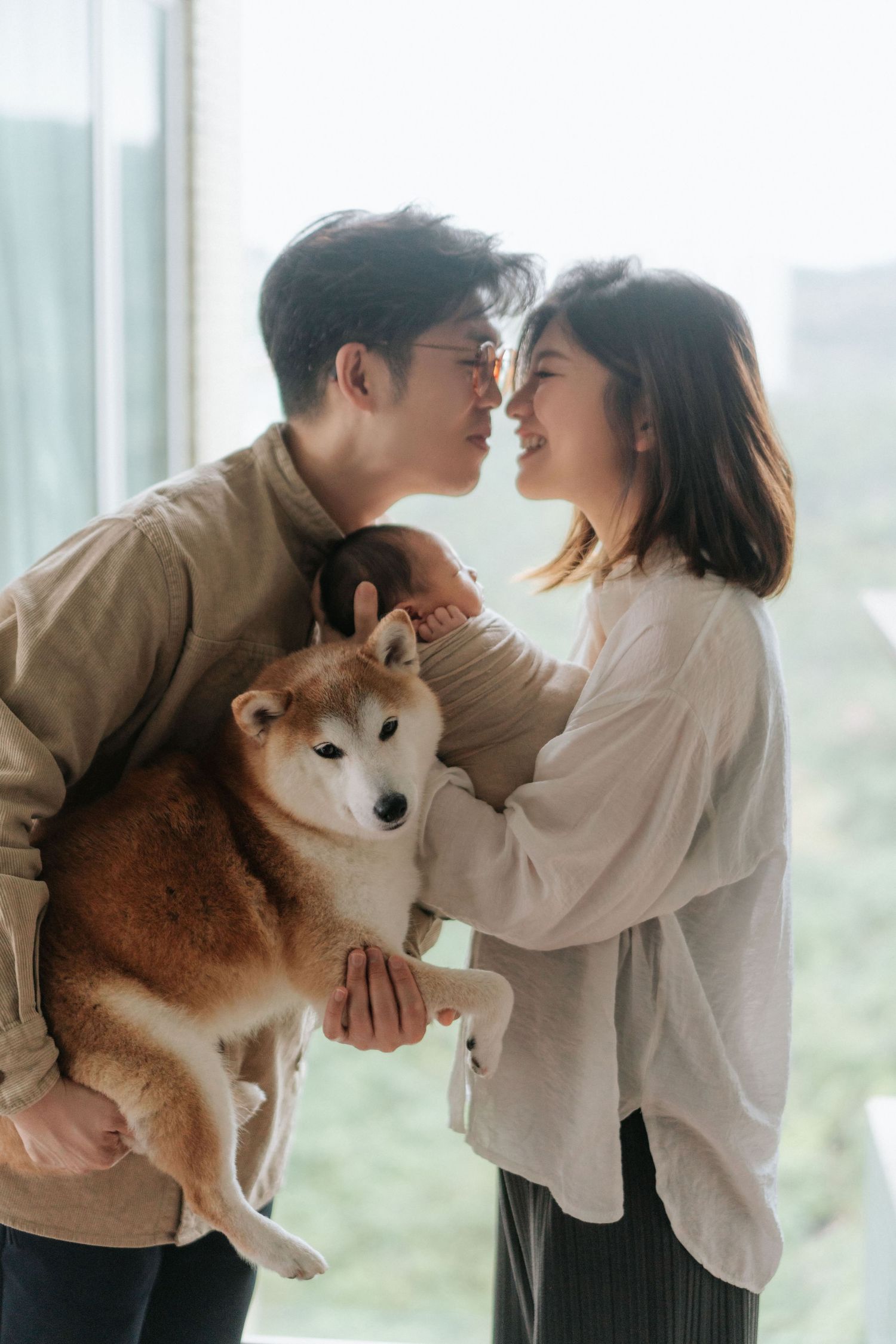 A couple shares an intimate moment while holding their Shiba Inu dog by a bright window.
