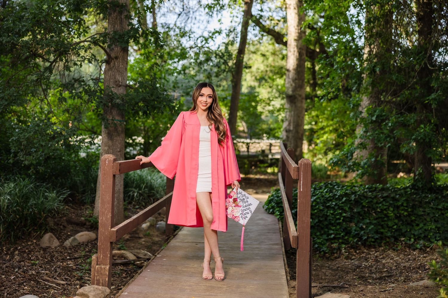 A woman in a flowing pink coat stands gracefully on a wooden bridge path surrounded by lush greenery.