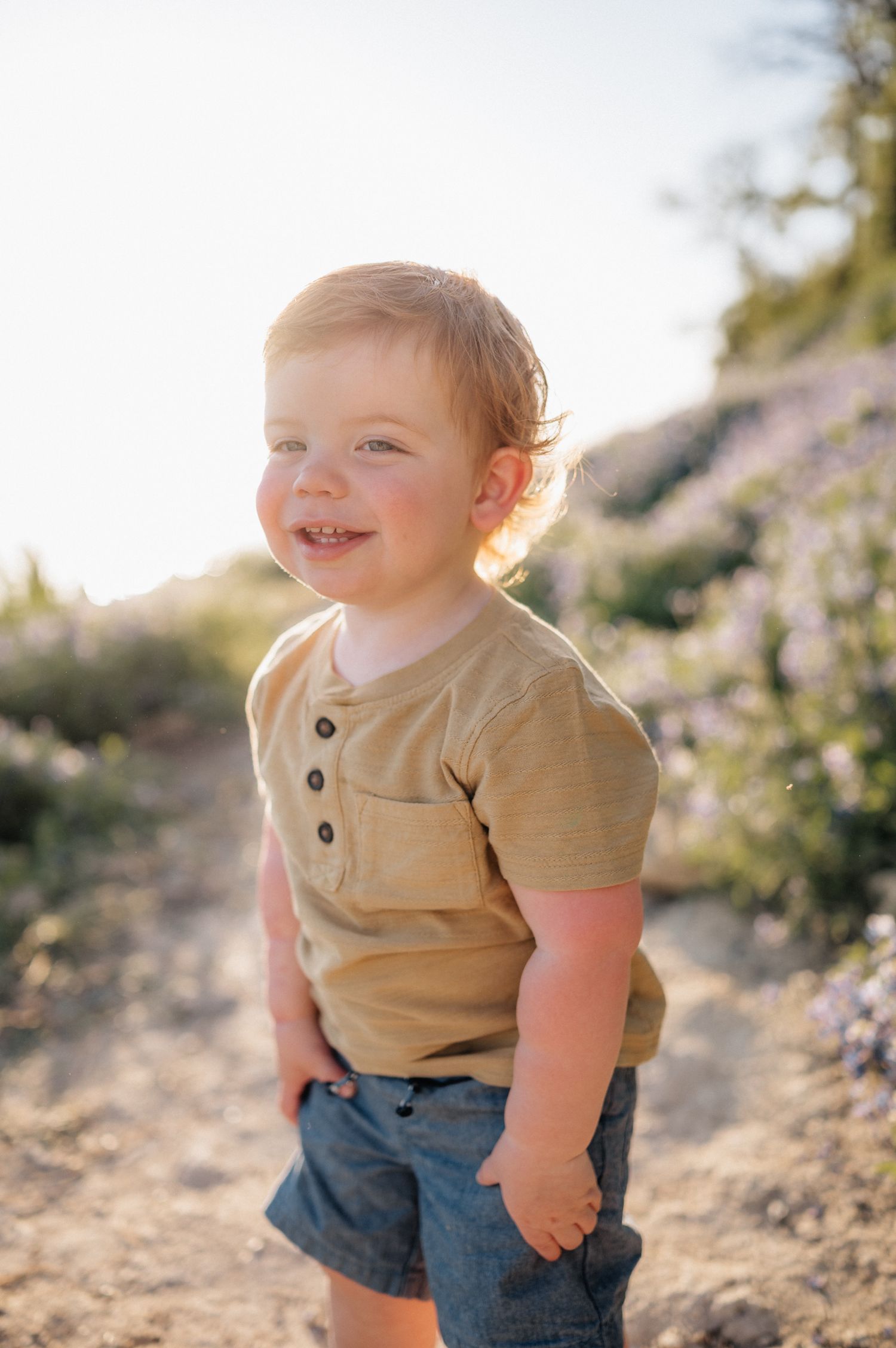 Toddler in tan shirt smiles while standing on dirt path among purple flowers.