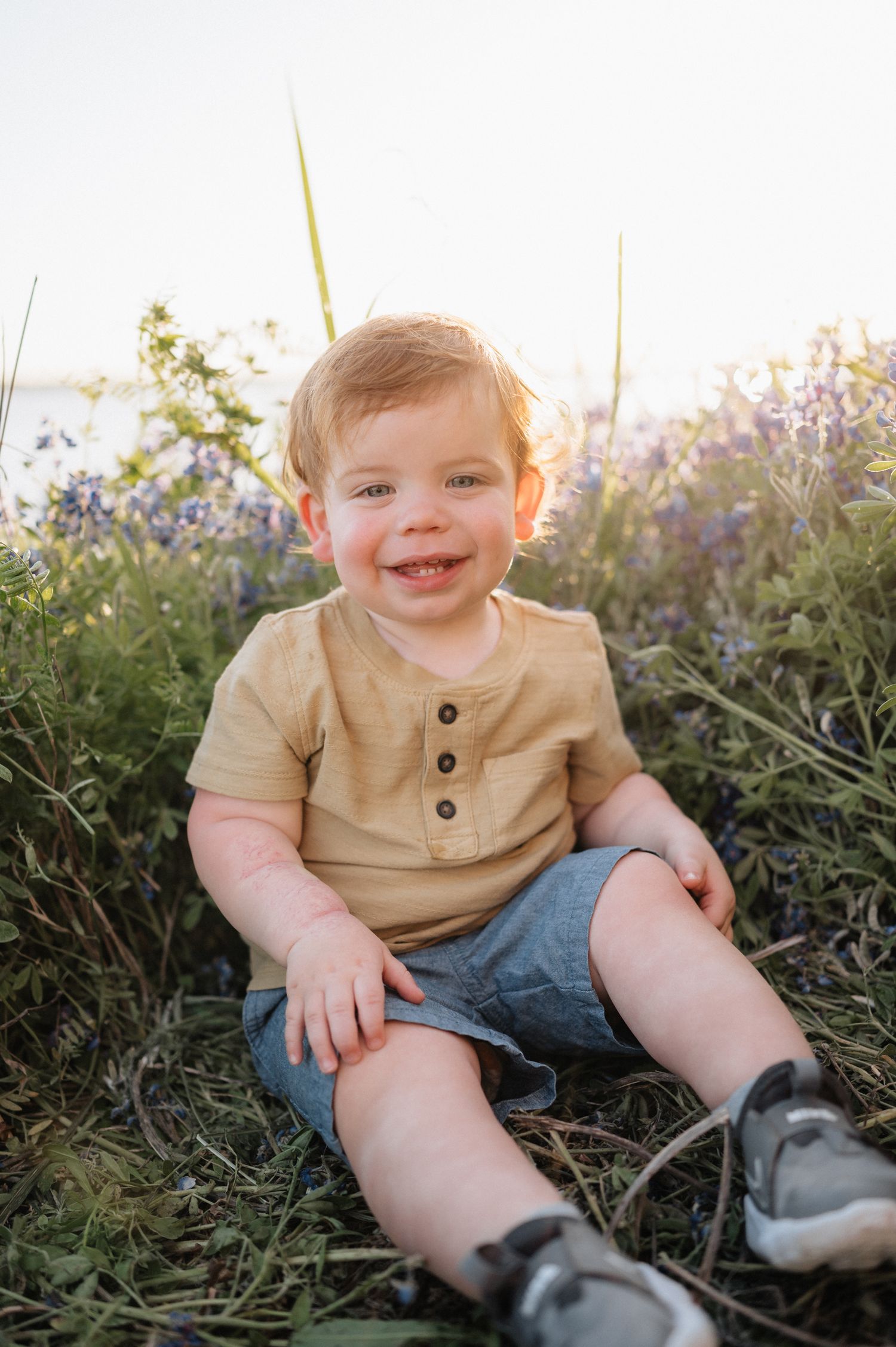 Toddler in tan henley shirt sits smiling among wildflowers at sunset.
