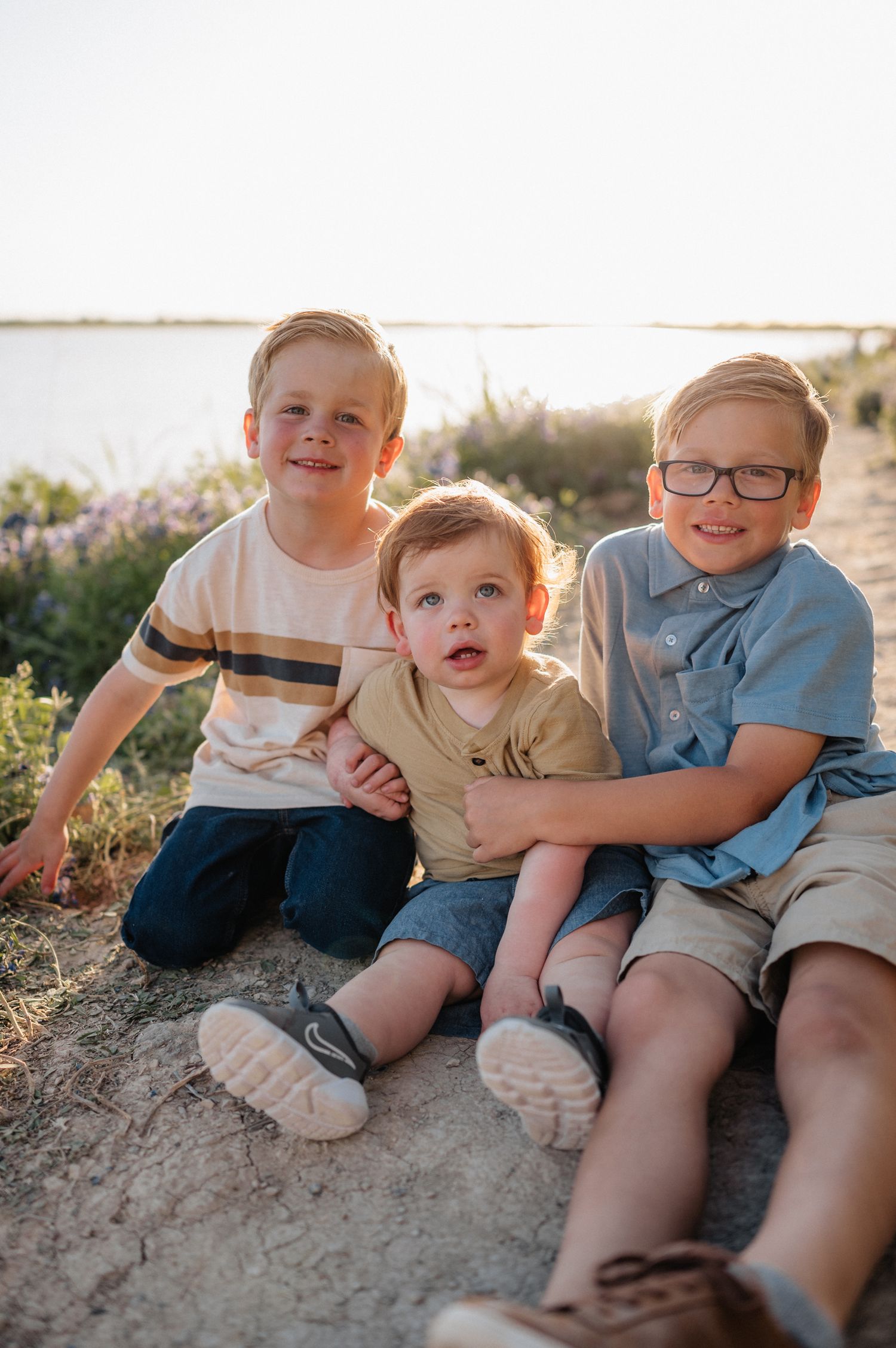 Three children sit together on dirt path with water and wildflowers in background.