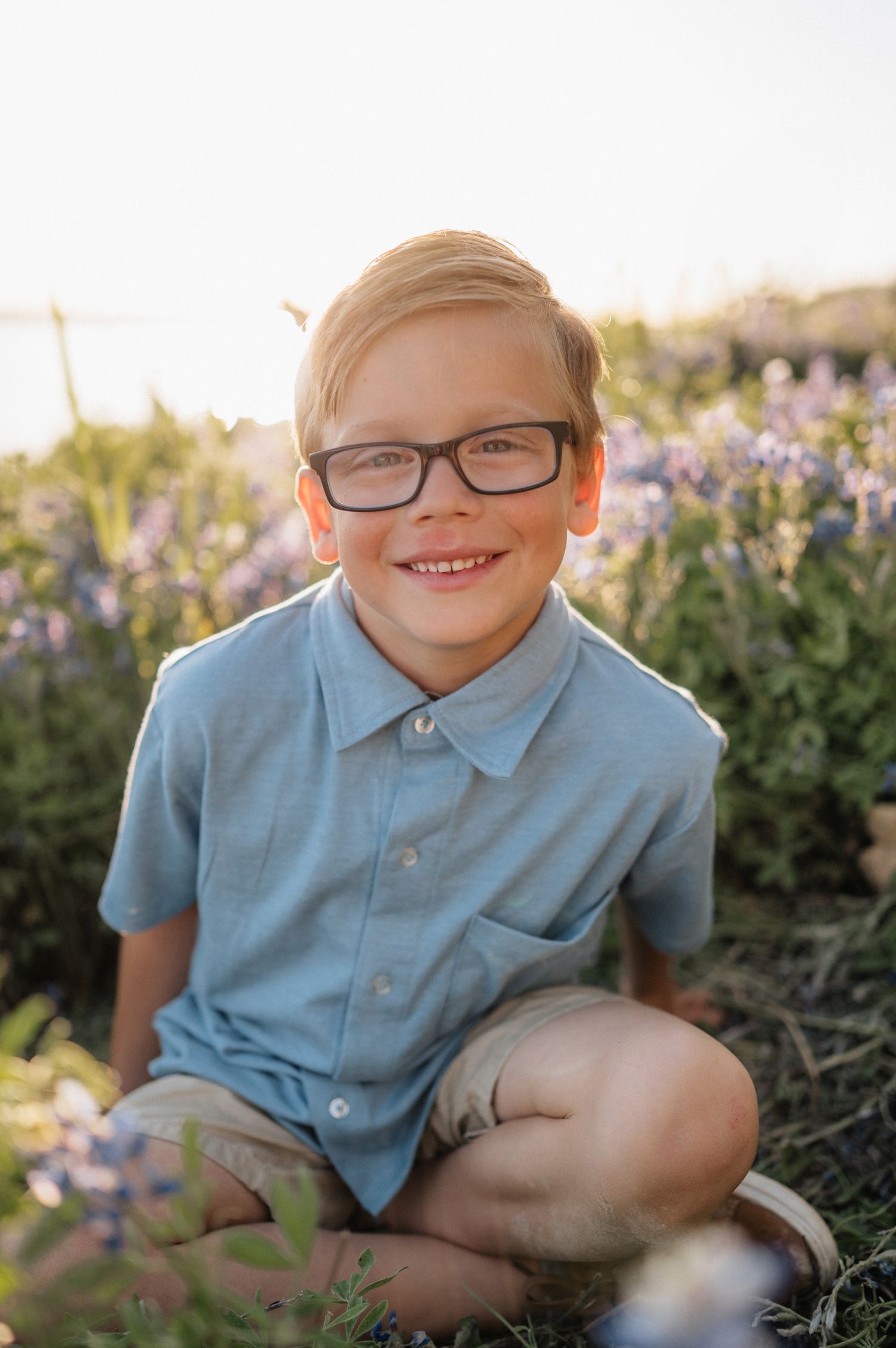 Young boy in blue shirt and glasses sits smiling in wildflower field during golden hour.