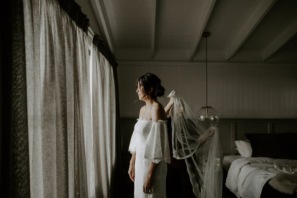 An elegant bride in an off-shoulder wedding dress stands by a window with flowing curtains in a moody interior setting.
