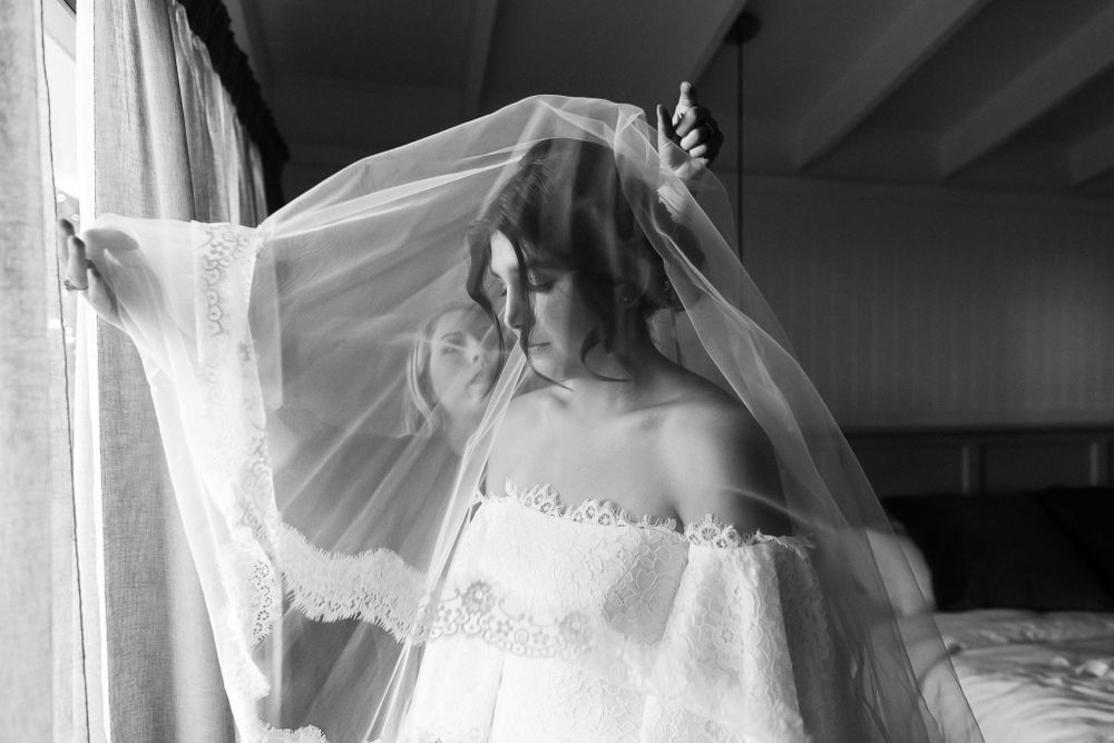 Black and white photo of a bride adjusting her delicate veil near a sunlit window.