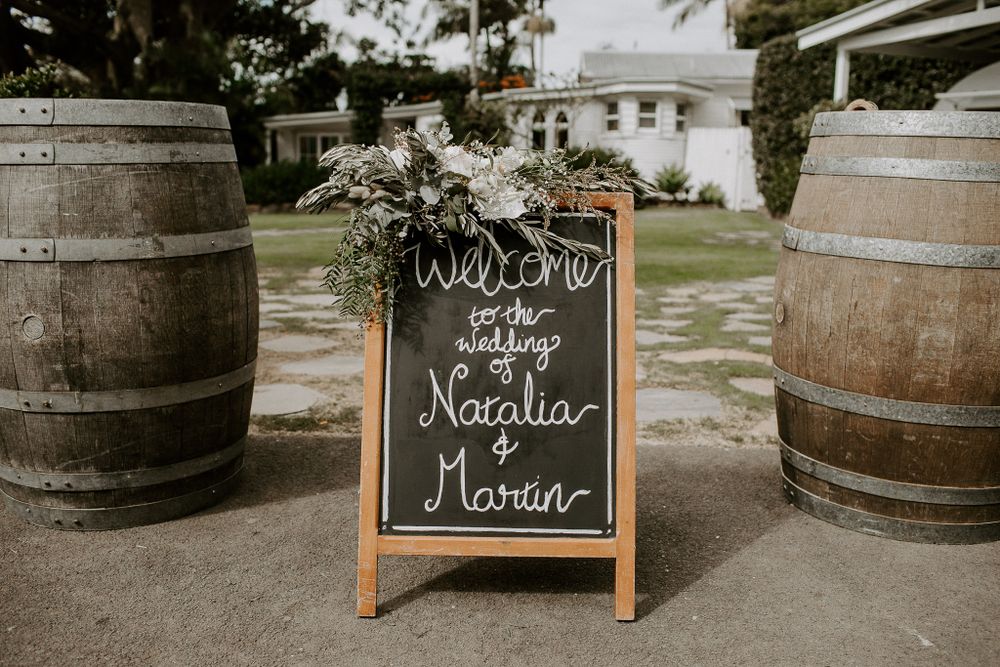 A rustic wedding welcome sign with names displayed between two wooden barrels outdoors.