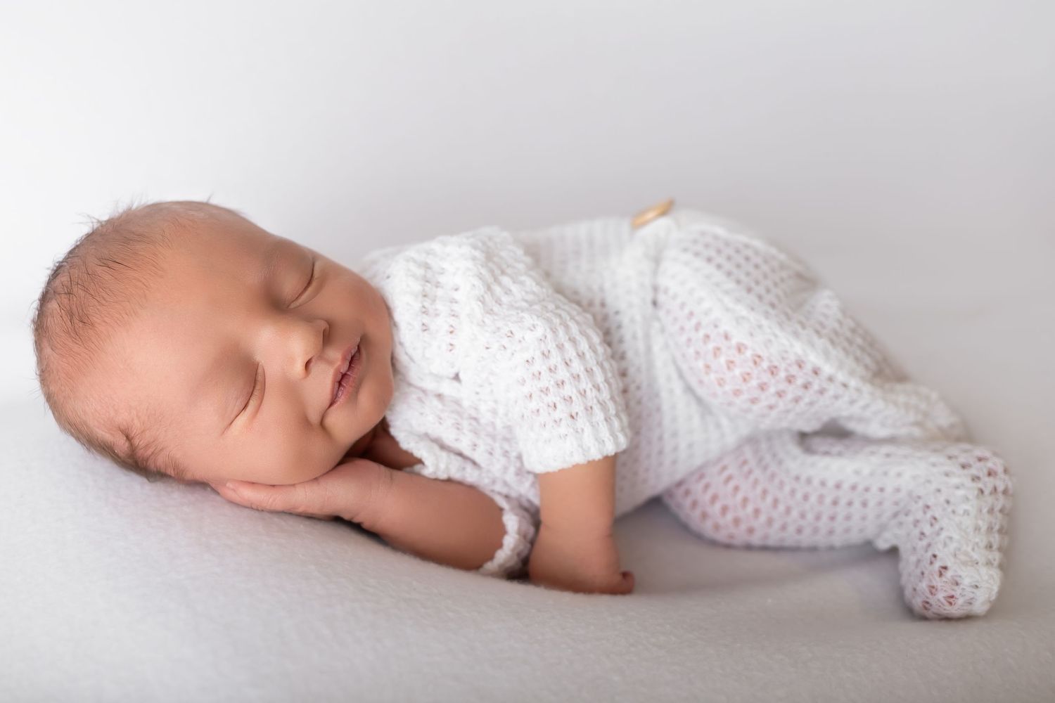 Sleeping newborn baby wearing white knitted outfit on light gray background.