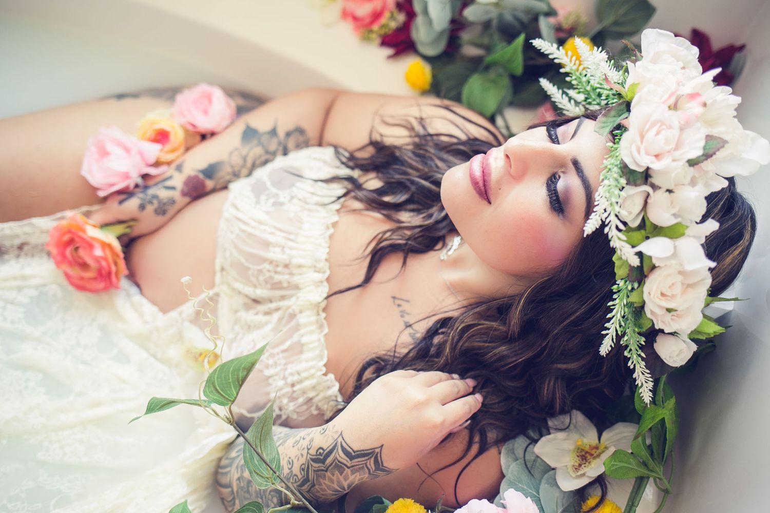 Tattooed woman in white lace gown lying among colorful flowers wearing a floral crown.