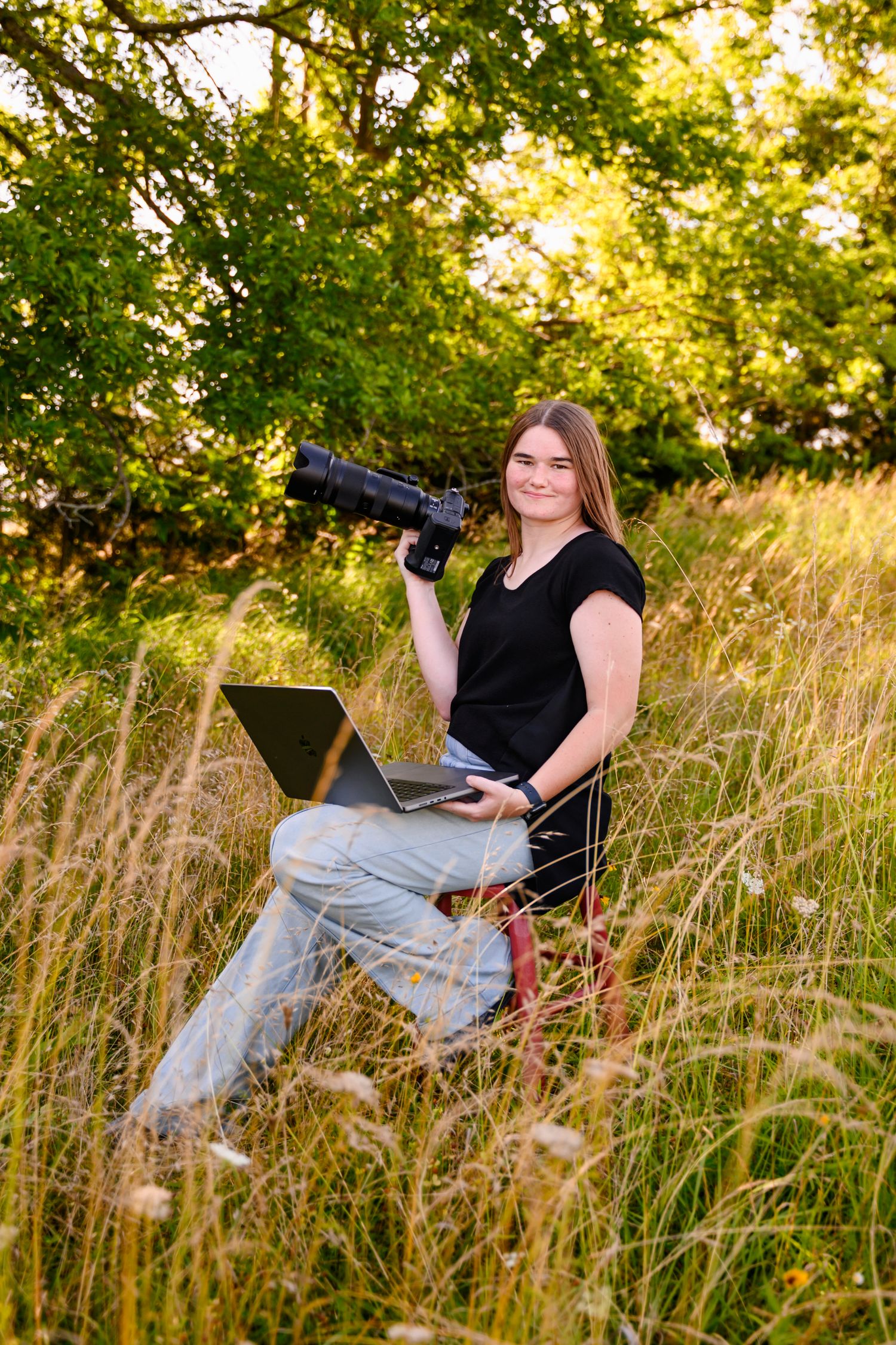 Professional photographer with camera and laptop sits in golden summer field during sunset.