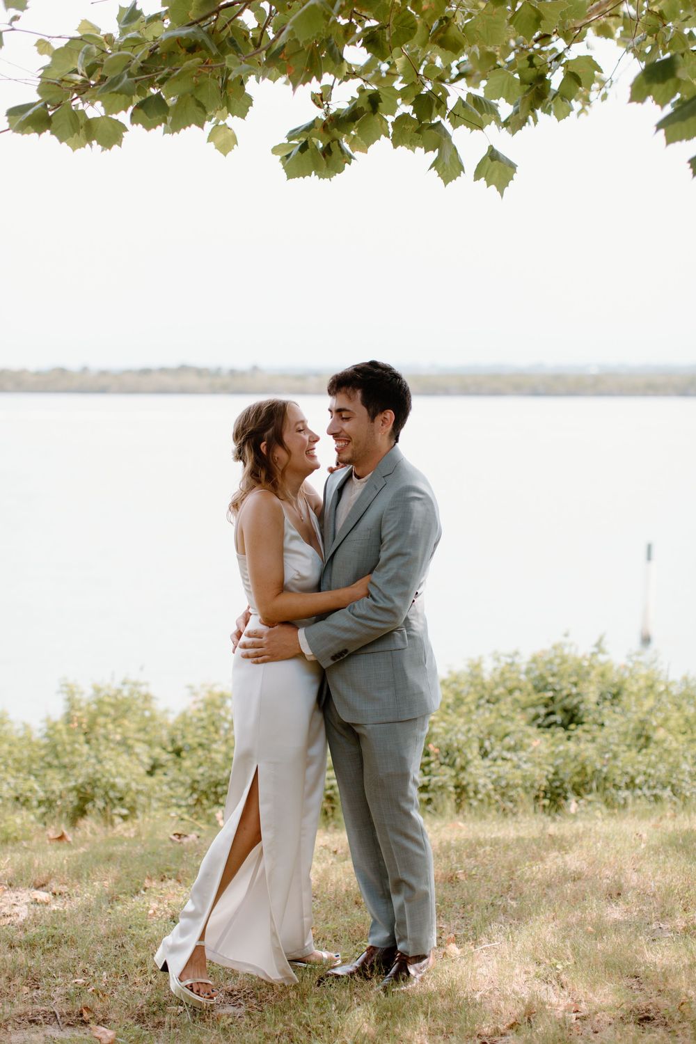 Couple embraces on hillside overlooking water in elegant wedding attire.