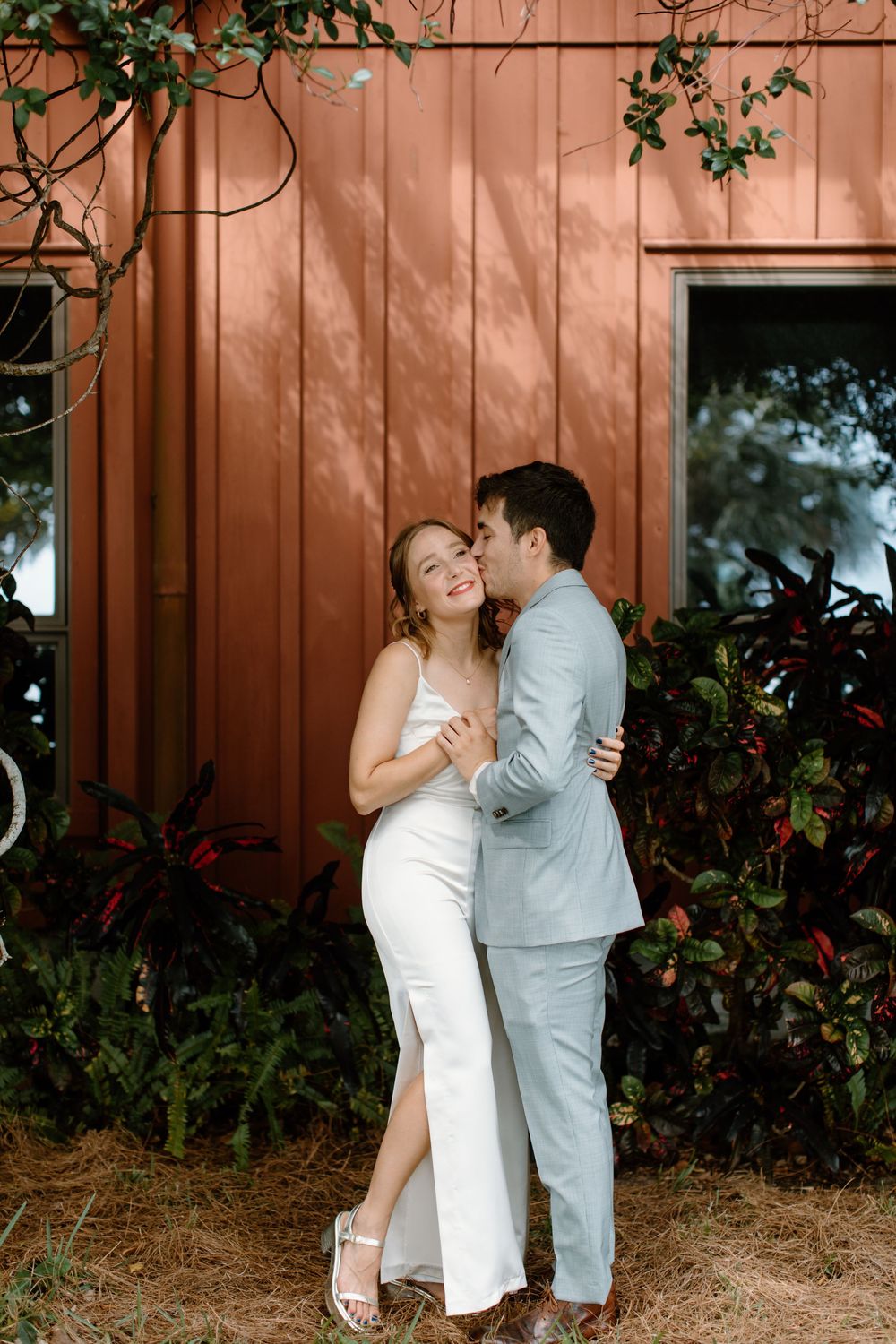 Couple shares intimate moment against coral colored barn wall with climbing vines.