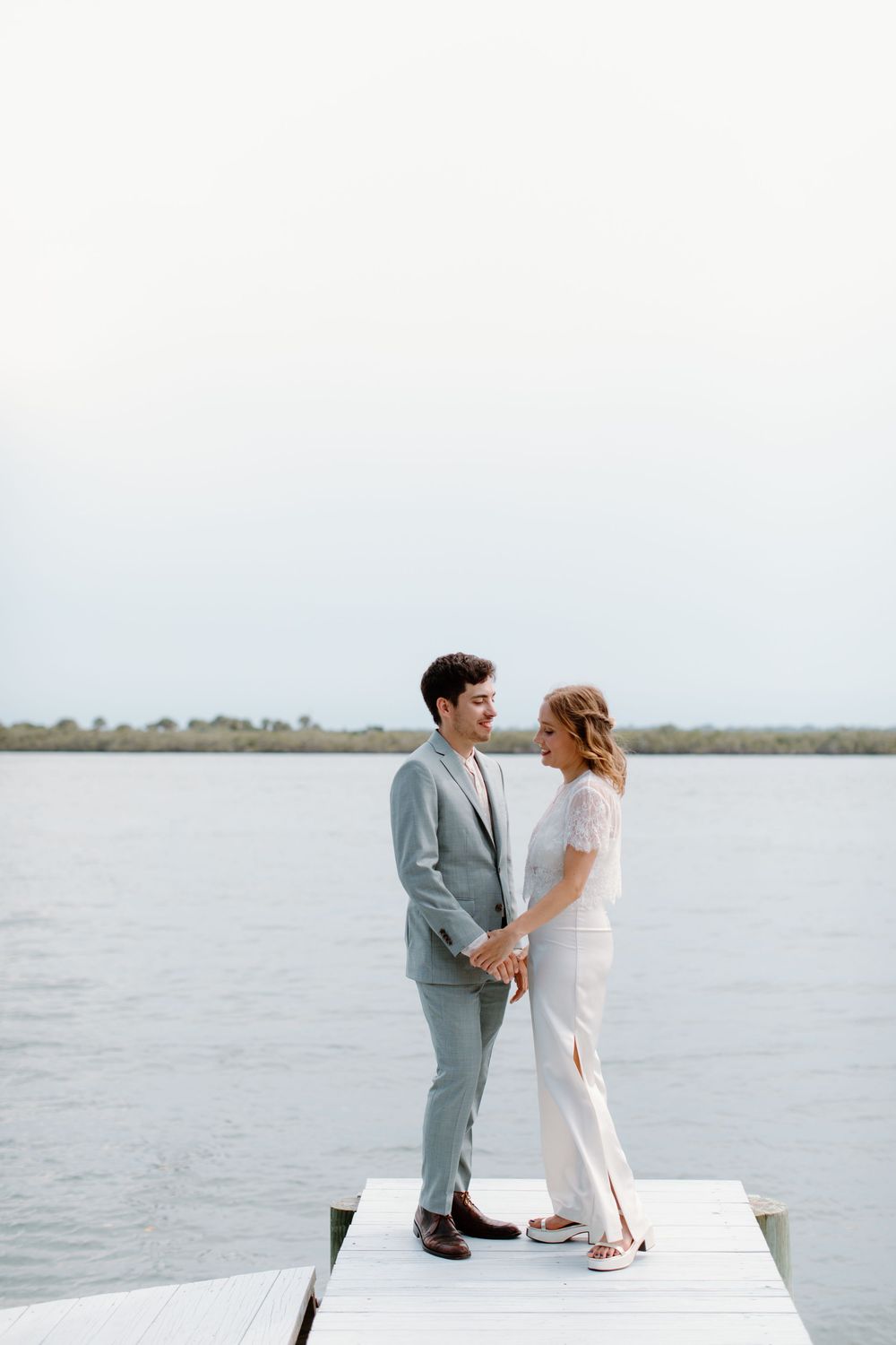 A romantic couple stands on a waterfront dock holding hands, with the groom in a gray suit and bride in white.